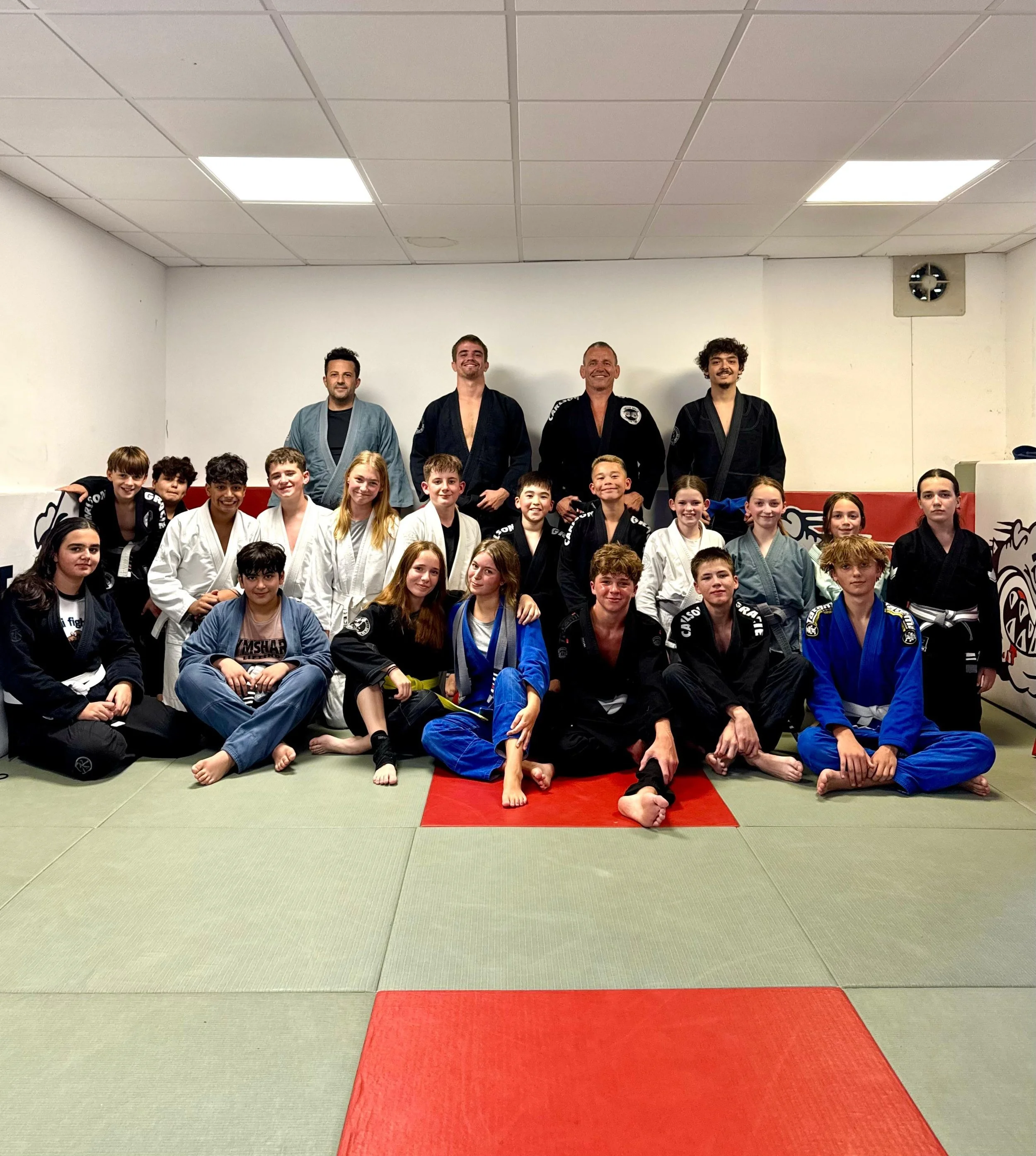 Group of children and adults in martial arts uniforms posing for a photo in a dojo. Several are sitting on or kneeling next to a red mat, while others stand behind them. The ceiling has fluorescent lights and a ventilation fan.