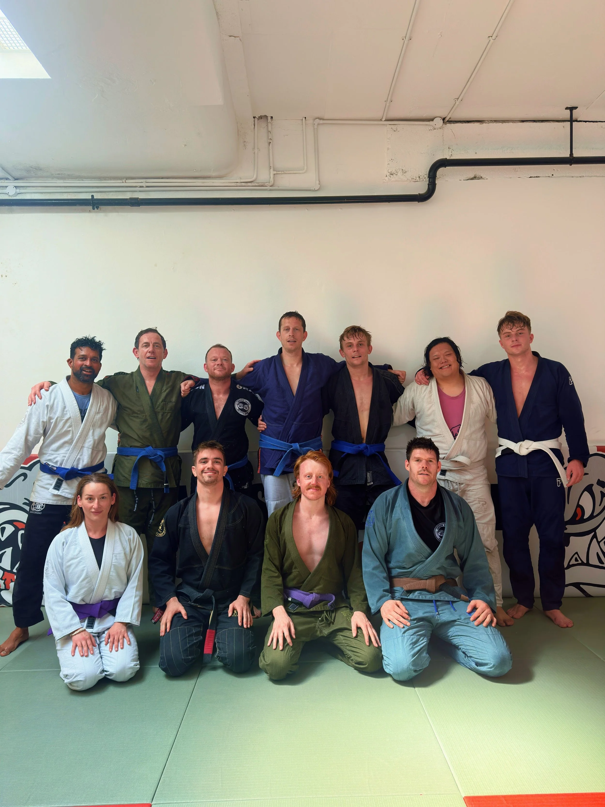 Group of eight martial artists in gis, standing and kneeling on mats in a dojo, posing for a photo.