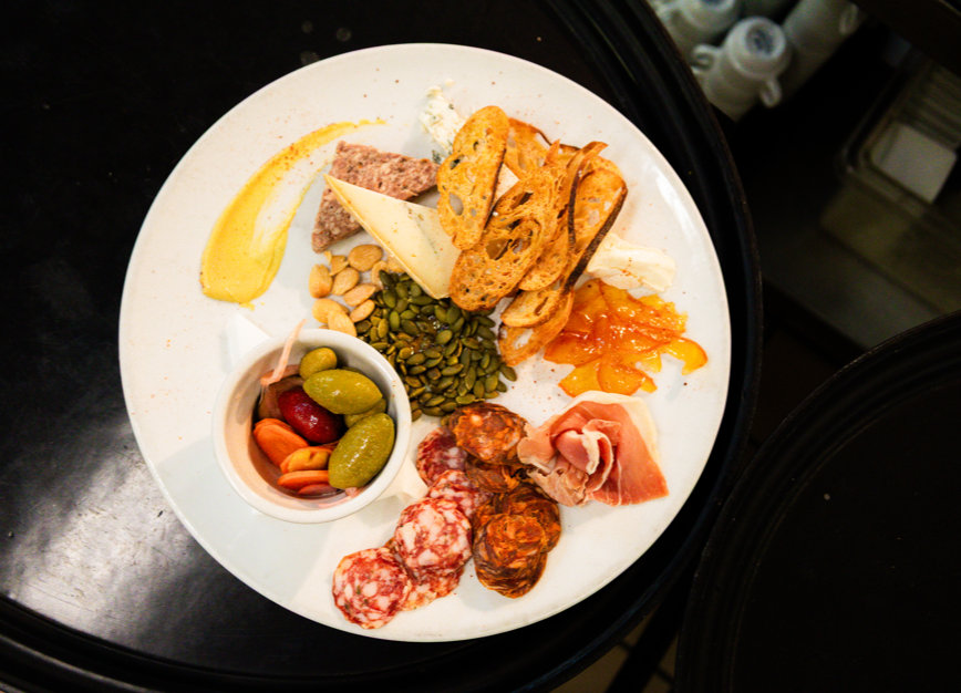 A plate with assorted snacks including sliced cured meats, cheese, crackers, assorted nuts, pickles, dried fruit, and spreads.