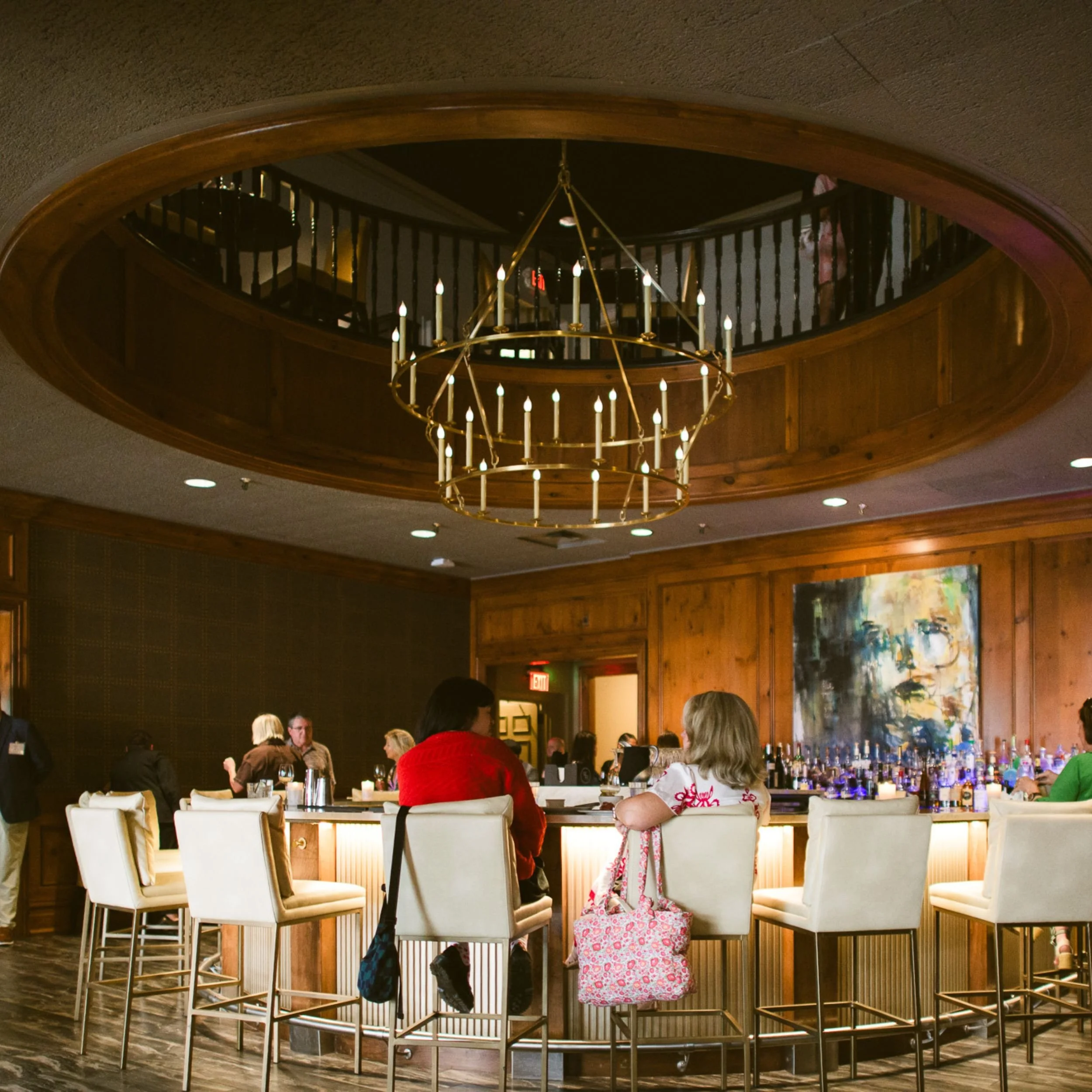 Interior of a restaurant or bar with people sitting at tables and a prominent round bar height table surrounded by chairs. There is a large, circular chandelier hanging from the ceiling and wood paneling on the walls.