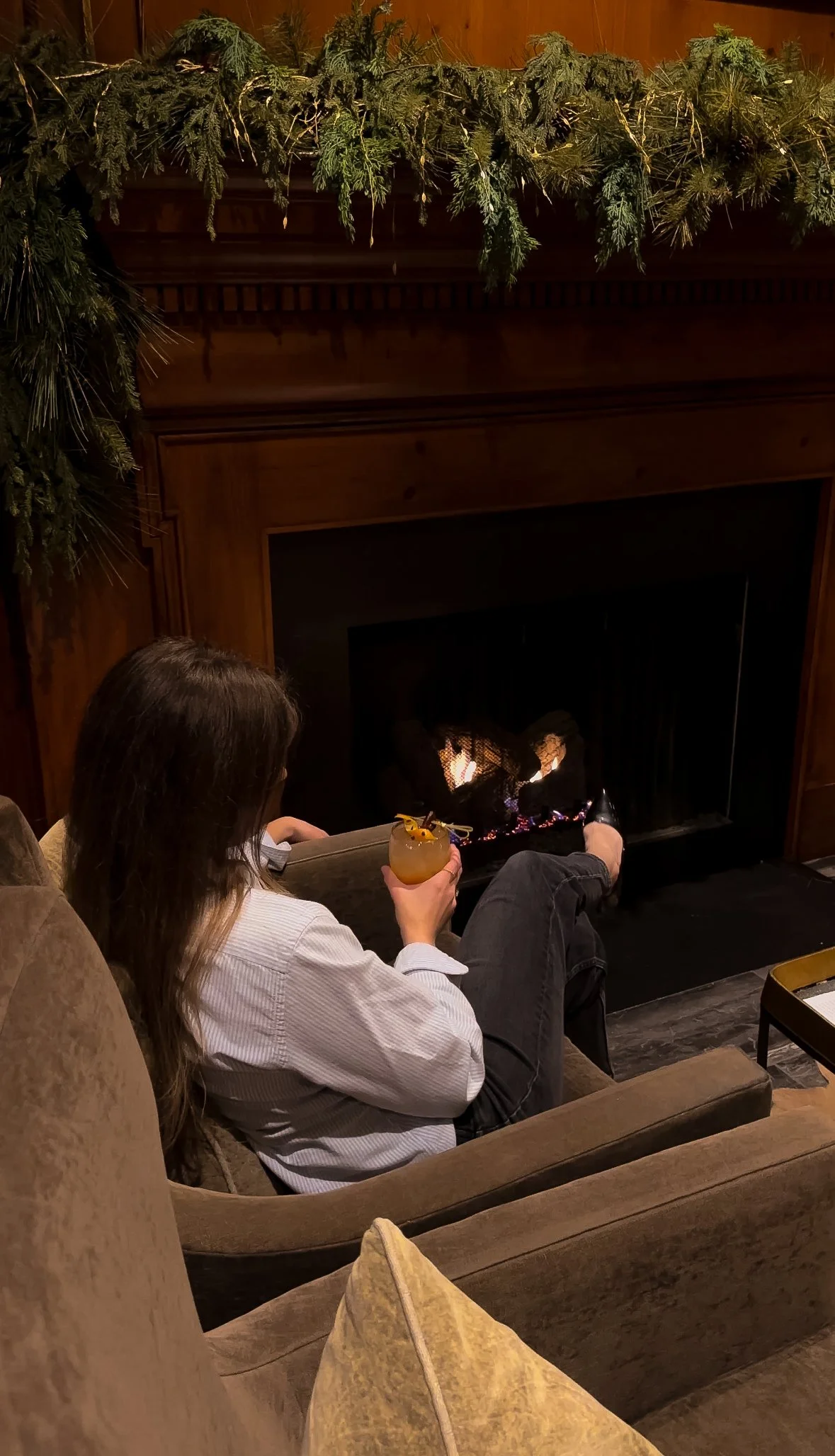 A woman with long brown hair is sitting on a beige couch near a fireplace, holding a yellow cocktail with a small umbrella and a straw. She is wearing a white shirt and black pants. The fireplace is lit, and the mantel is decorated with greenery. The