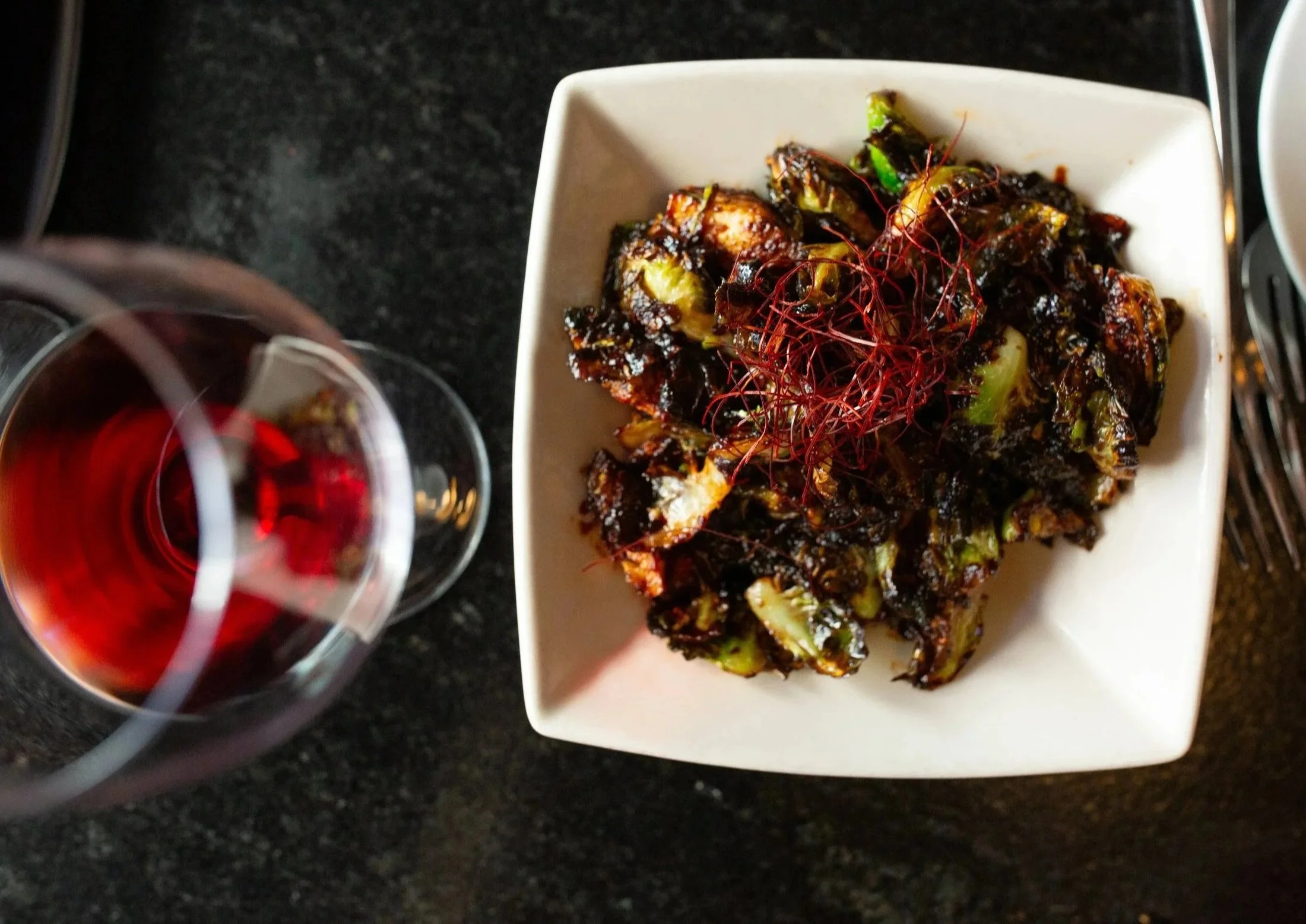 A white rectangular plate with roasted vegetables topped with red chili threads, next to a glass of red wine on a dark countertop.