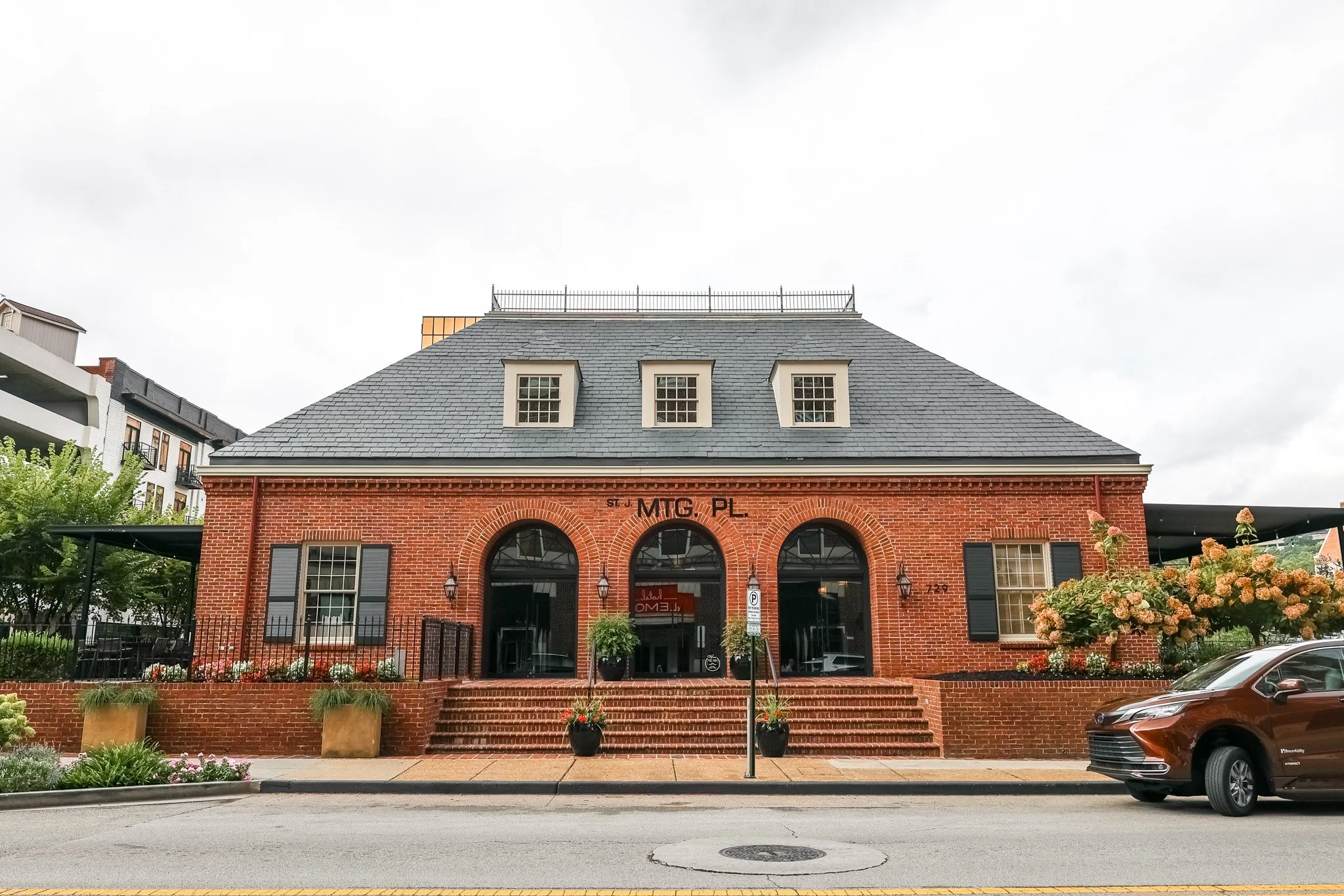 A brick building with large arched windows and black shutters, three dormer windows on the roof, and a sign that reads 'MIG PL.' There are stairs leading to the entrance and potted plants on either side. The building is surrounded by trees and parked cars.