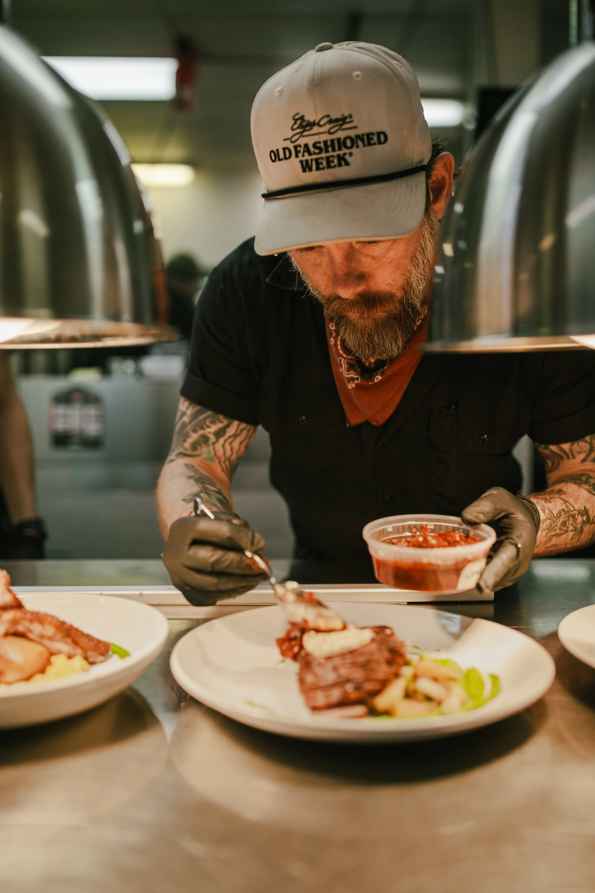 A man with tattoos and a beard, wearing a beige cap with text, preparing food behind a counter with plates of cooked meat and vegetables in front of him in a restaurant kitchen.