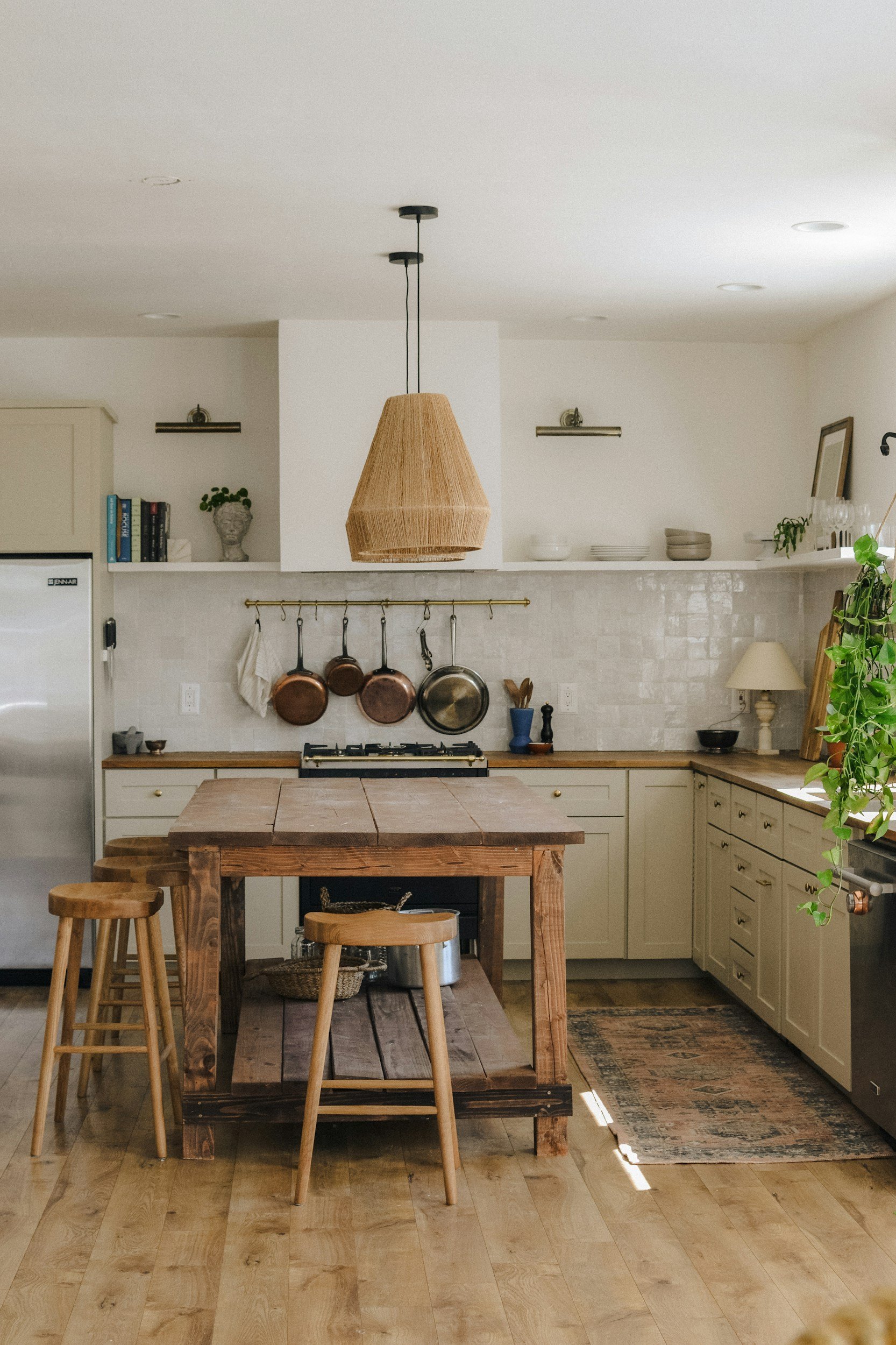 A rustic kitchen with a wooden table and stools, beige cabinets, hanging copper pans, and open shelves with books, bowls, and plants. A woven lampshade hangs from the ceiling.