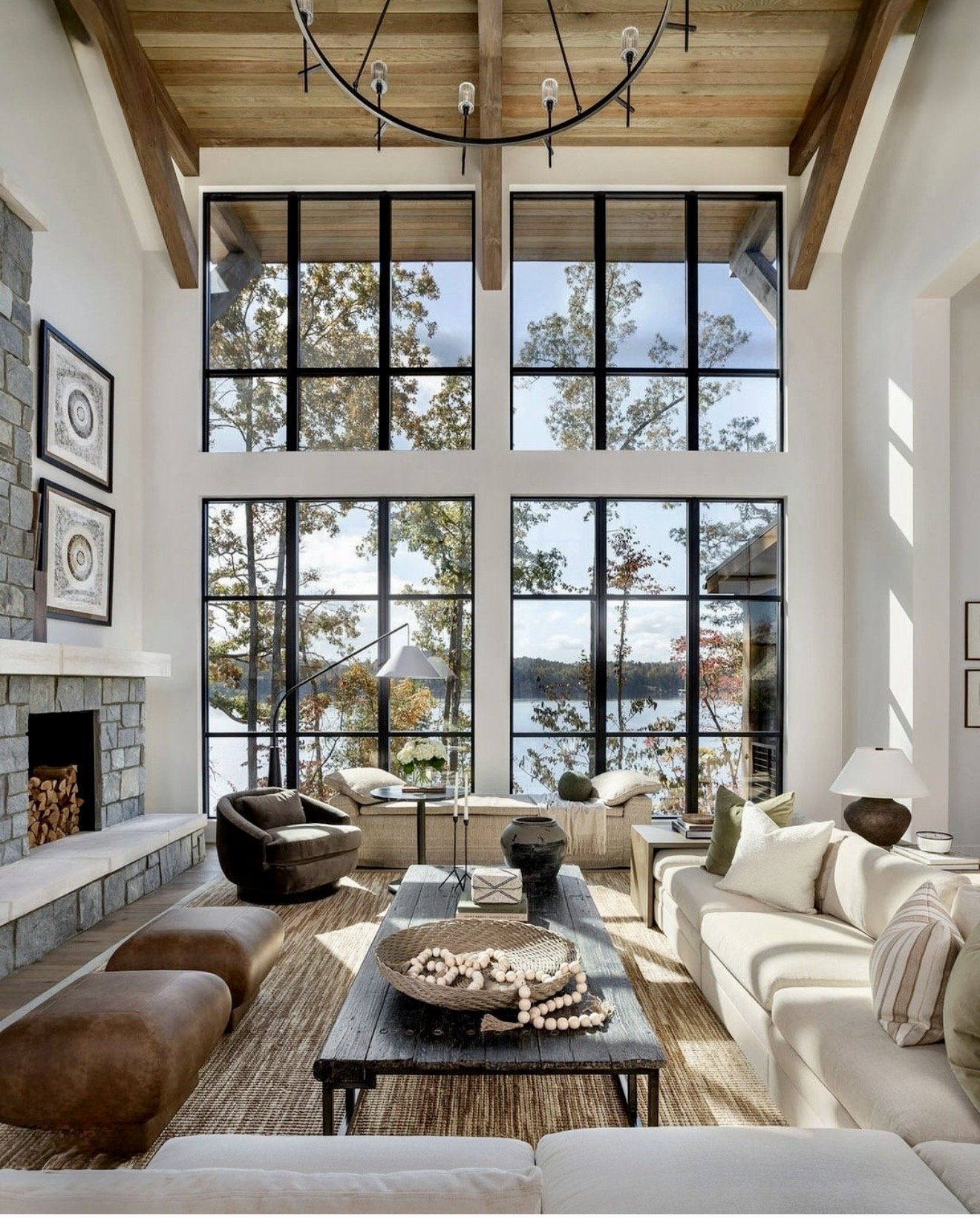 A spacious living room with large grid-style windows overlooking a view of trees and water. The room features a stone fireplace, cozy seating including a white sofa, chairs, and ottomans, and a rustic wooden coffee table with decorative items. The ceiling is made of wood with exposed beams and a large round chandelier hanging from it.