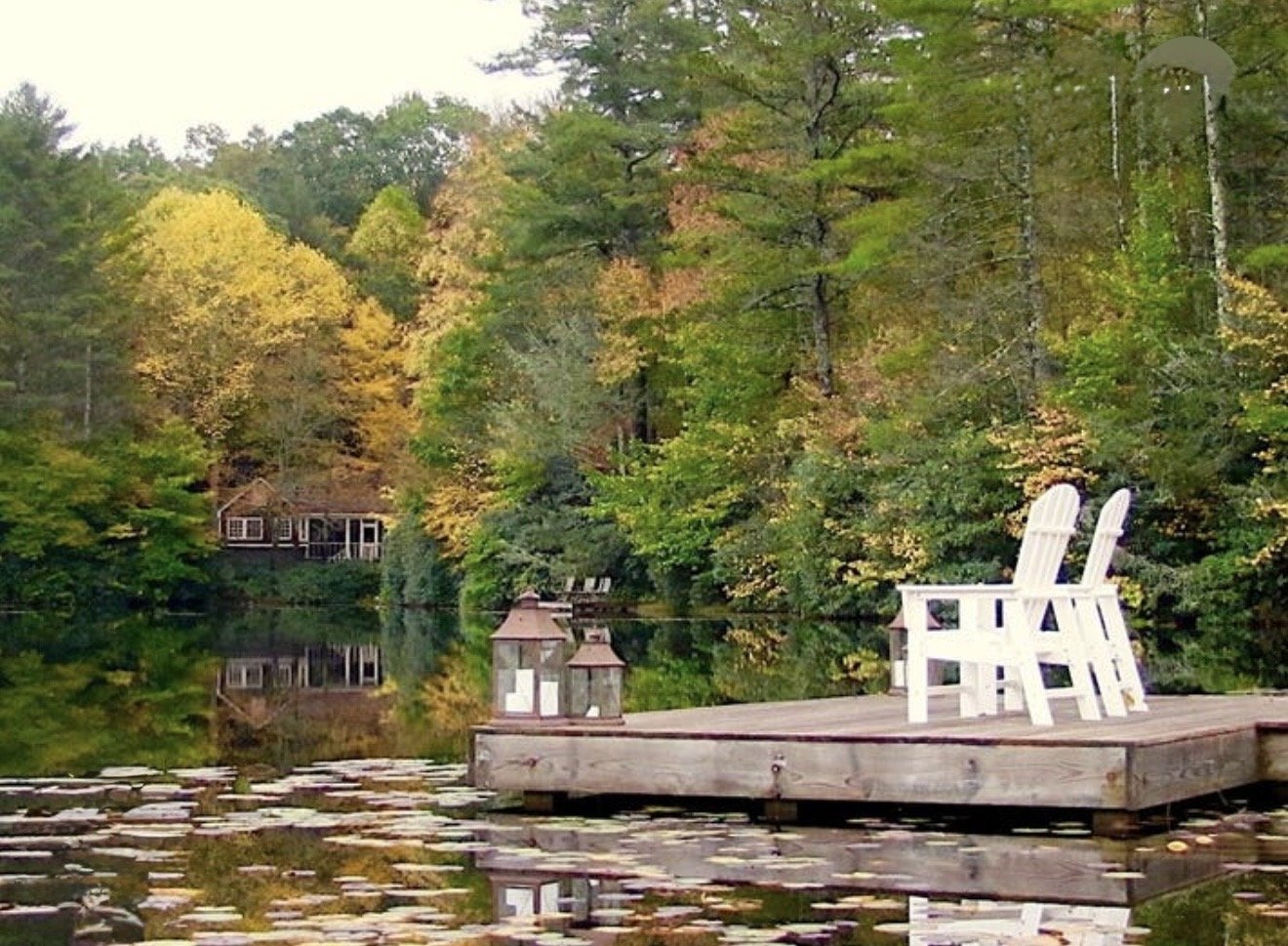 Two white Adirondack chairs on a wooden dock overlooking a calm lake surrounded by colorful trees with autumn foliage.
