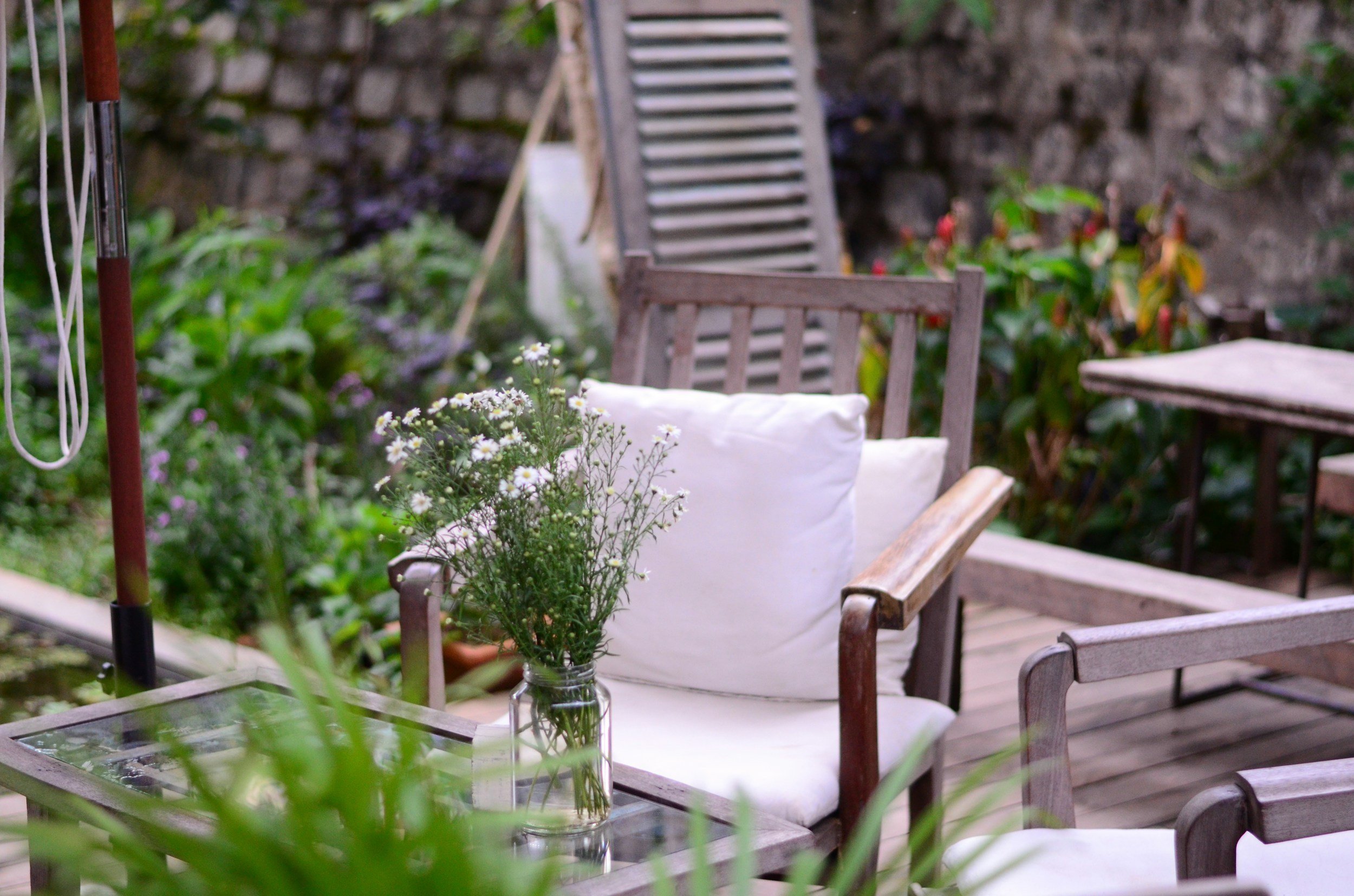 Outdoor patio with wooden chairs, a glass table with a vase of white and purple flowers, surrounded by plants and greenery, with a stone wall in the background.