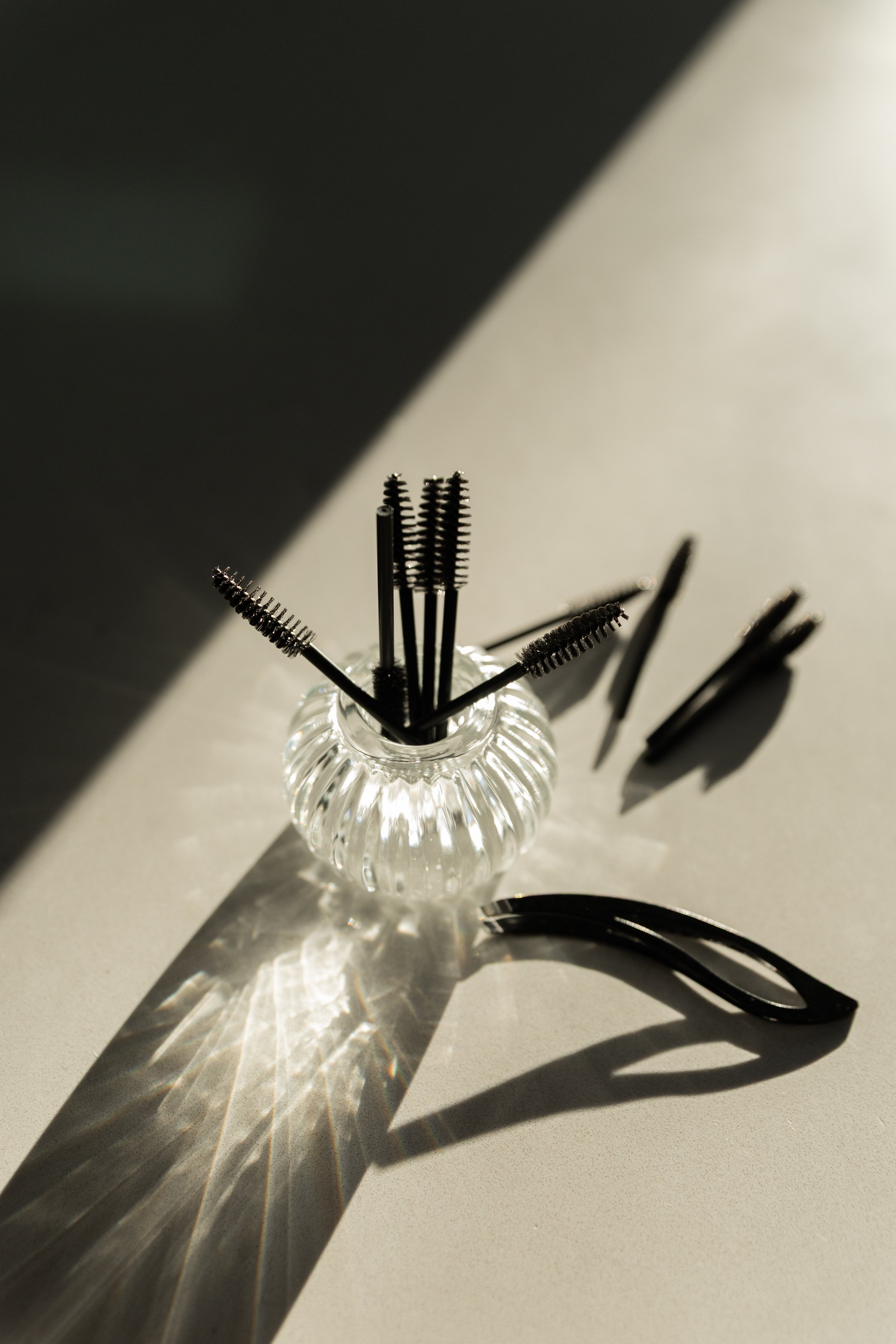 A small glass vase holding black eyelashes cleaning tools casting a shadow and reflections on a white surface with shadow dividing the image into light and dark areas.
