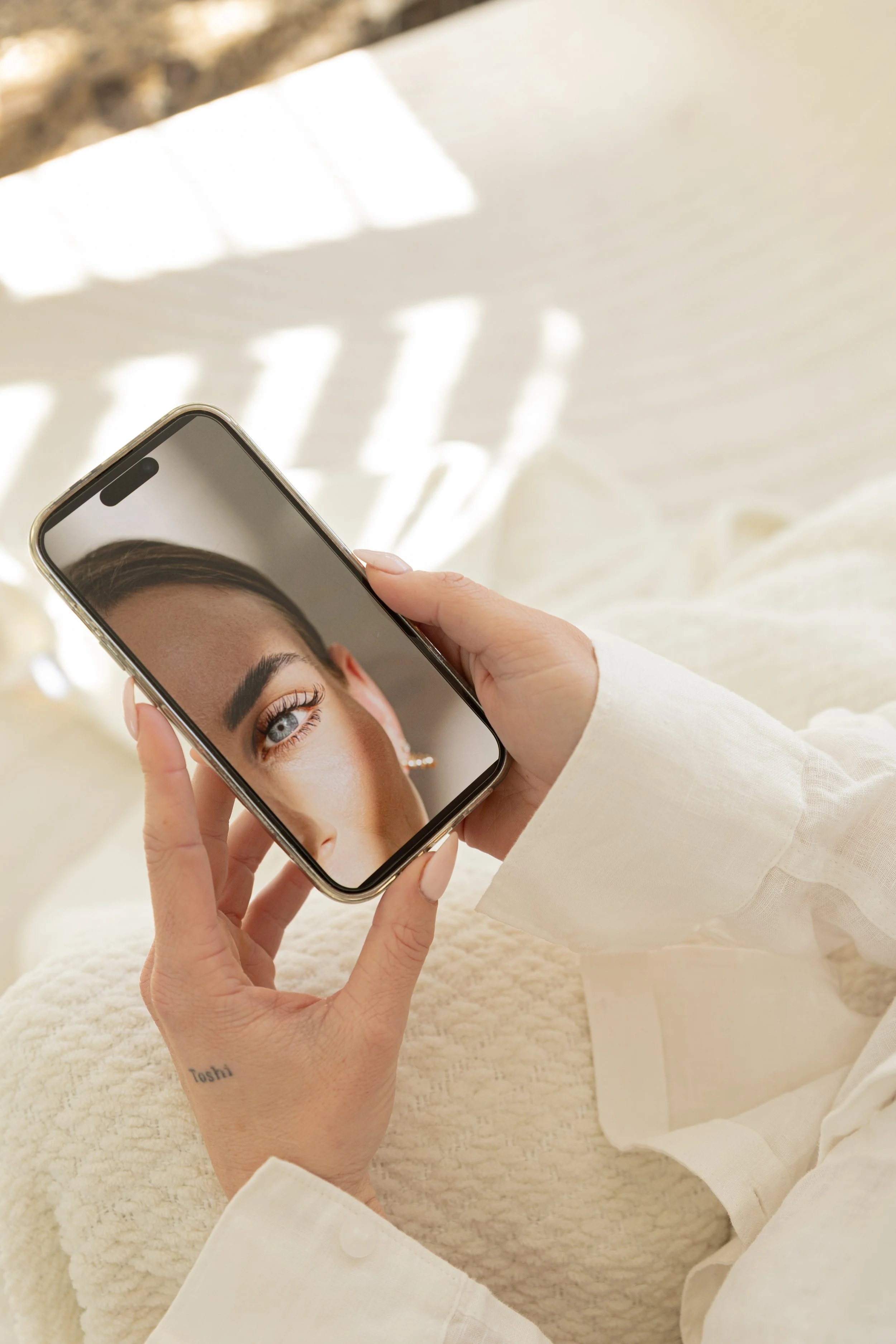 Person holding a smartphone with a photo of a woman with bold makeup and eyebrows displayed on the screen. The person has a tattoo on their wrist.