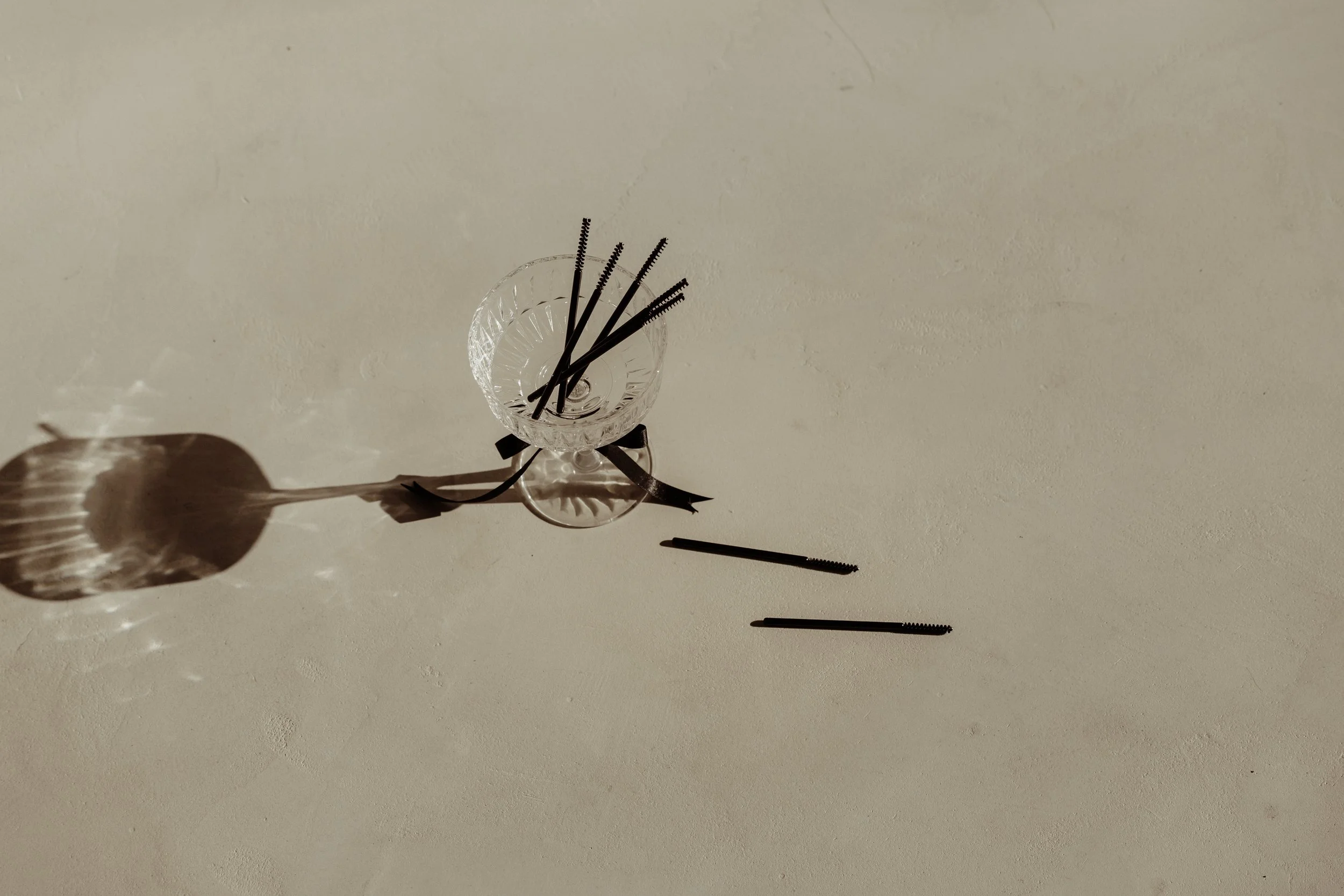 Several black hairpins arranged in and around a small glass bowl, some scattered on a light-colored surface. The bowl has a decorative pattern and a black ribbon tied around its base. The light casts shadows of the hairpins and bowl on the surface.