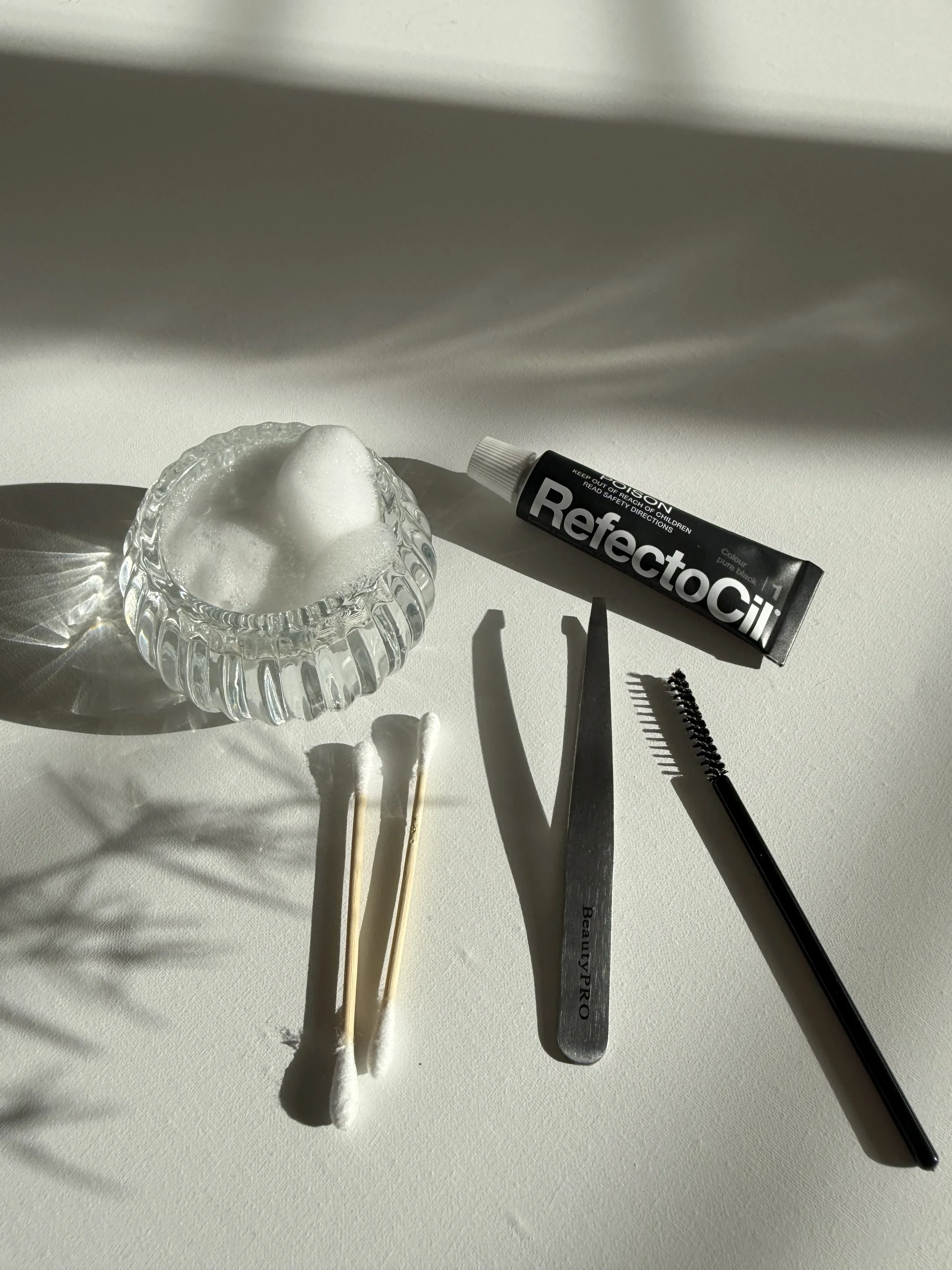 A glass bowl with a dollop of white foam, three cotton swabs, a pair of scissors, a leather spatula, and a mascara brush on a white surface, with shadows cast by natural light.