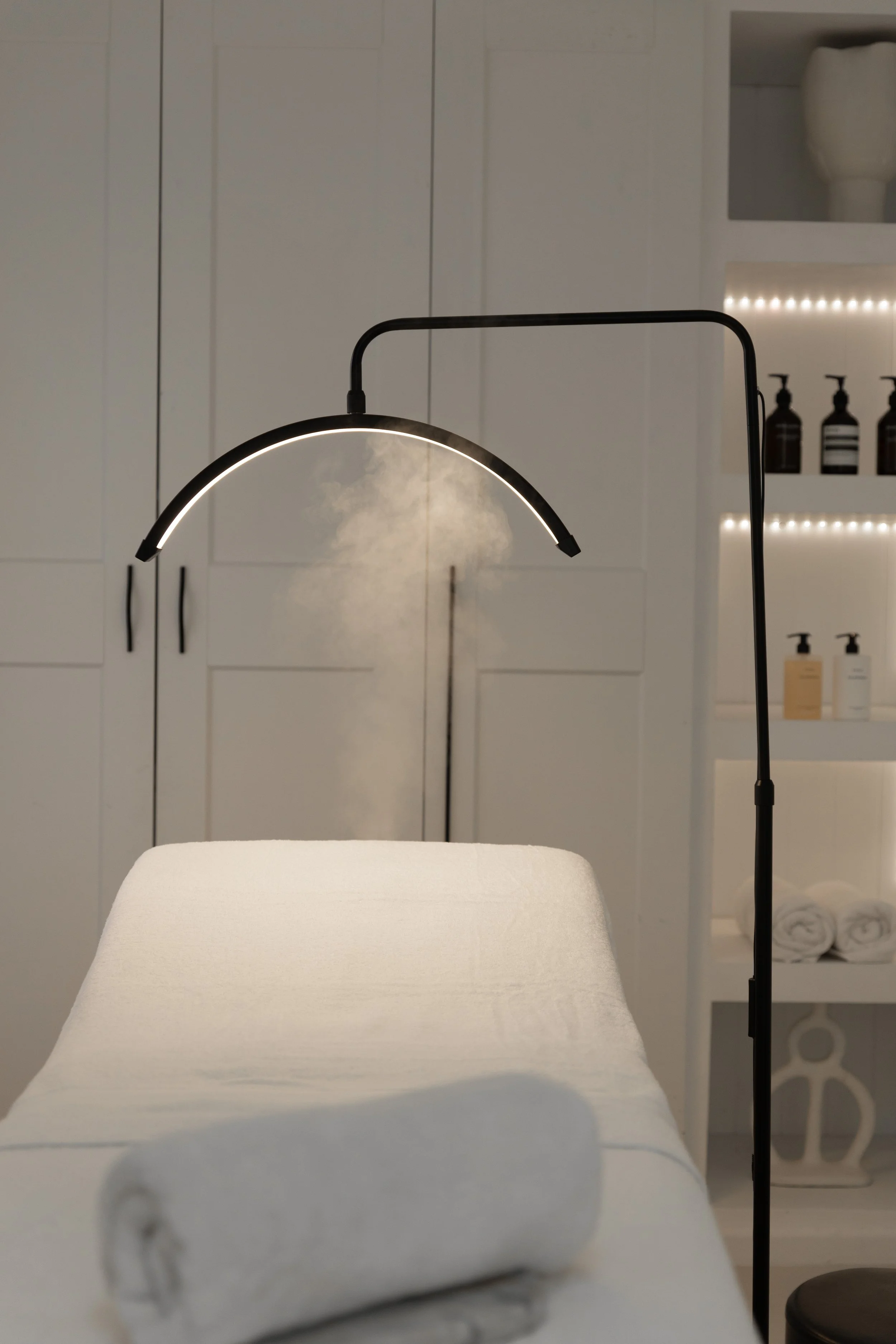 A white table or bed with a neatly folded white towel in the foreground, a modern black floor lamp with a curved light above, and a white cabinet with black handles and several bottles and towels in the background, suggesting a spa or wellness setting.