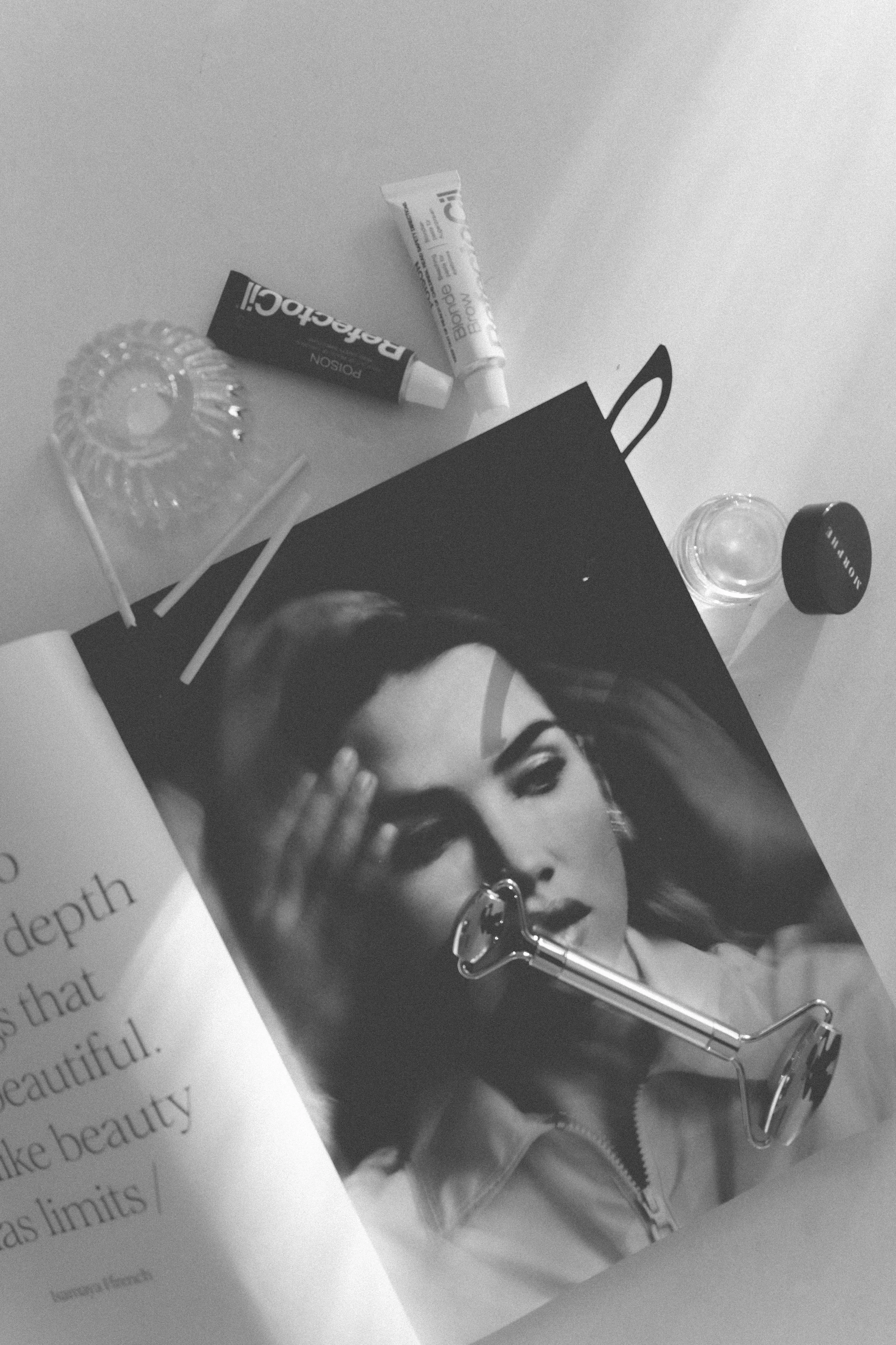Black and white photo of a magazine page with a woman with dark hair, bright lipstick, and wearing glasses, holding her hand with nail polish near her face. A lipstick container, a small jar, a box, and a roller brush are on the magazine.