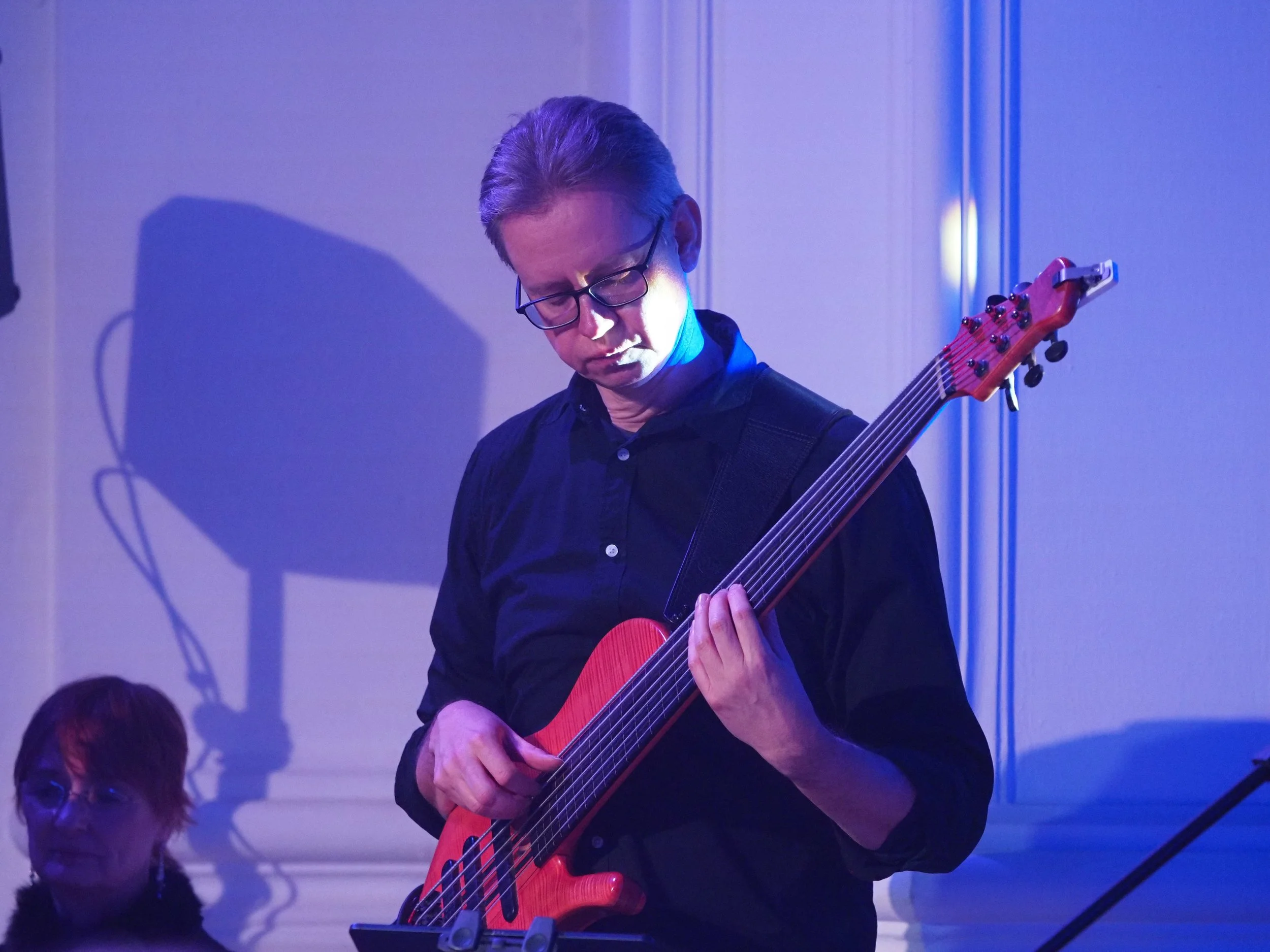Ein Mann mit Brille spielt Gitarre bei einem Musikauftritt in einem Raum mit blauer Beleuchtung. Ein Kind sitzt im Vordergrund und beobachtet.