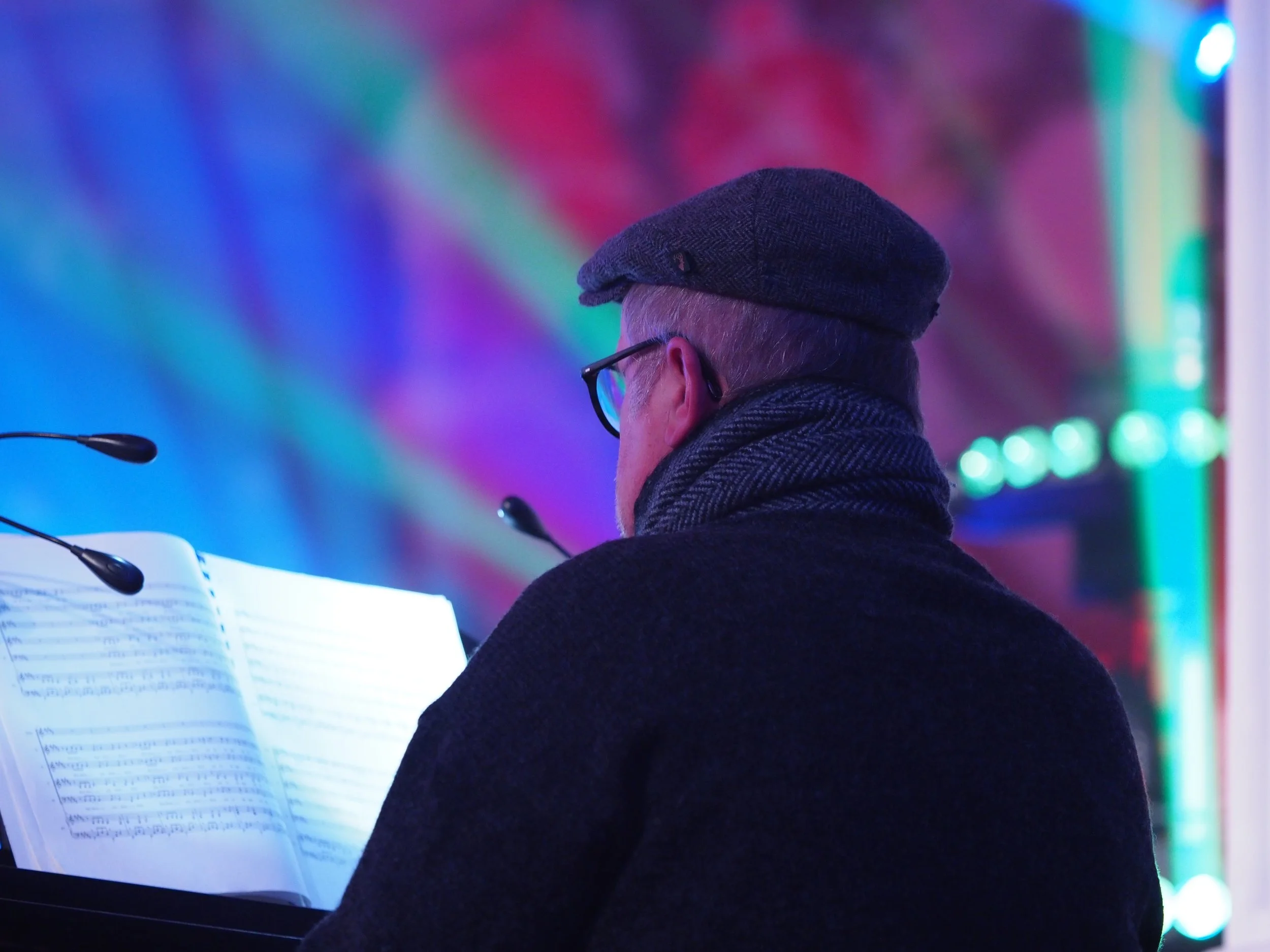 Ein älterer Mann trägt eine Mütze, eine Brille und einen Schal. Er sitzt an einem Mikrofon und liest ein Dokument mit Text. Im Hintergrund leuchten bunte, neonartige Lichter in Blau, Rot und Grün.