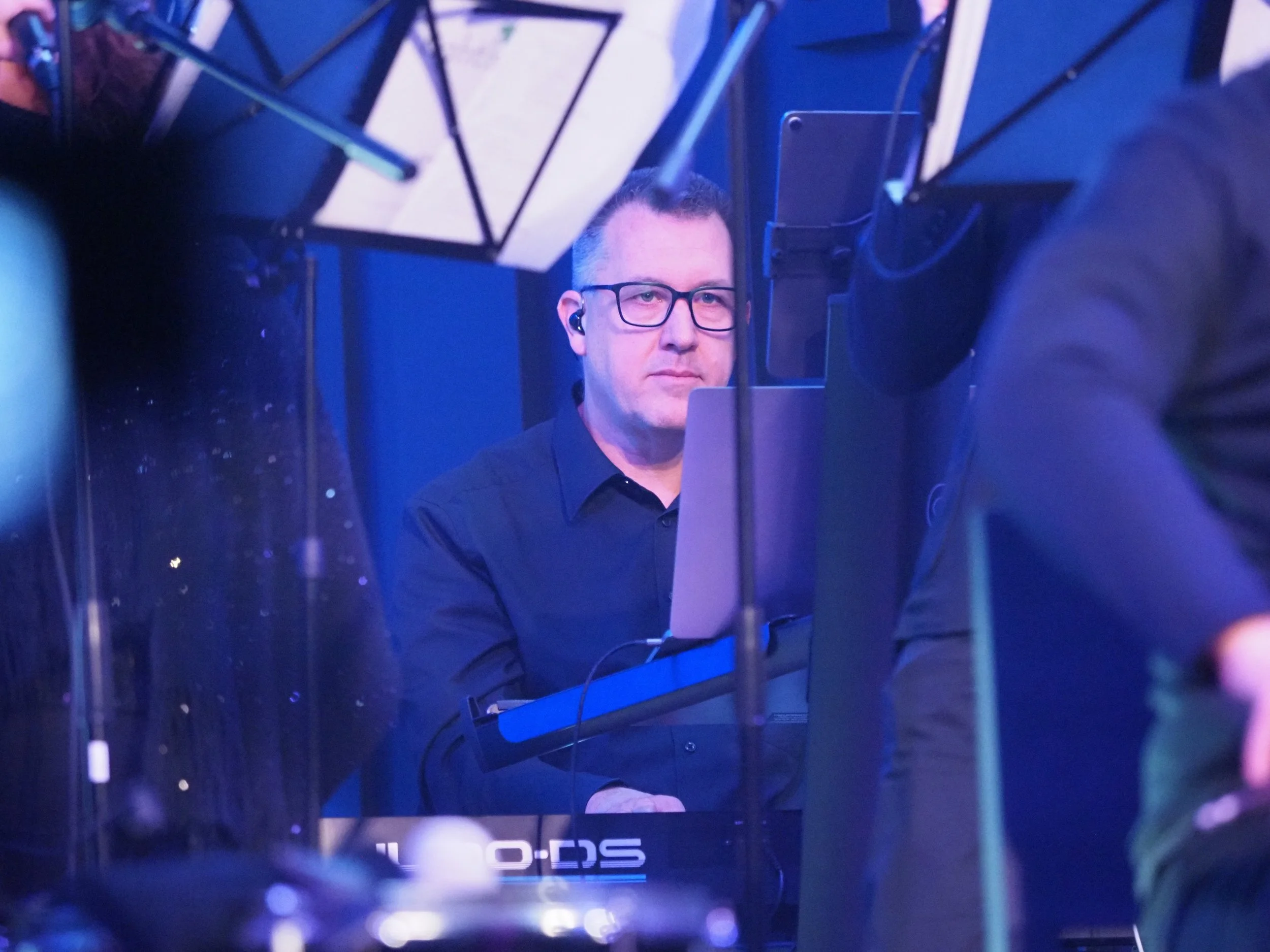 Mann mit Brille sitzt vor einem Laptop in einer dunklen, blaulichte Atmosphäre, umgeben von Schirmen und technischen Geräten.
