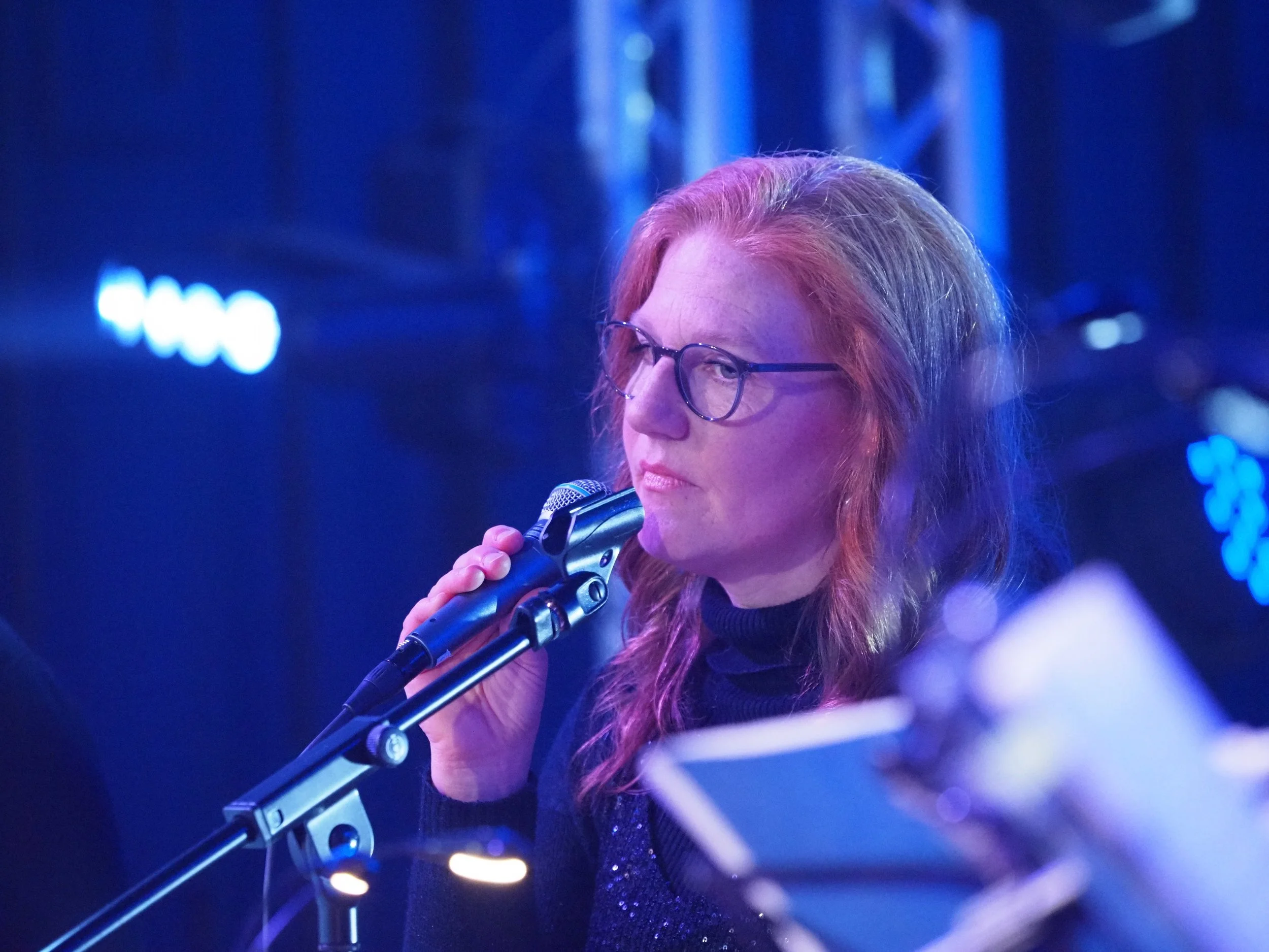 Frau mit roten Haaren und Brille singt in ein Mikrofon bei blauem Bühnenlicht.