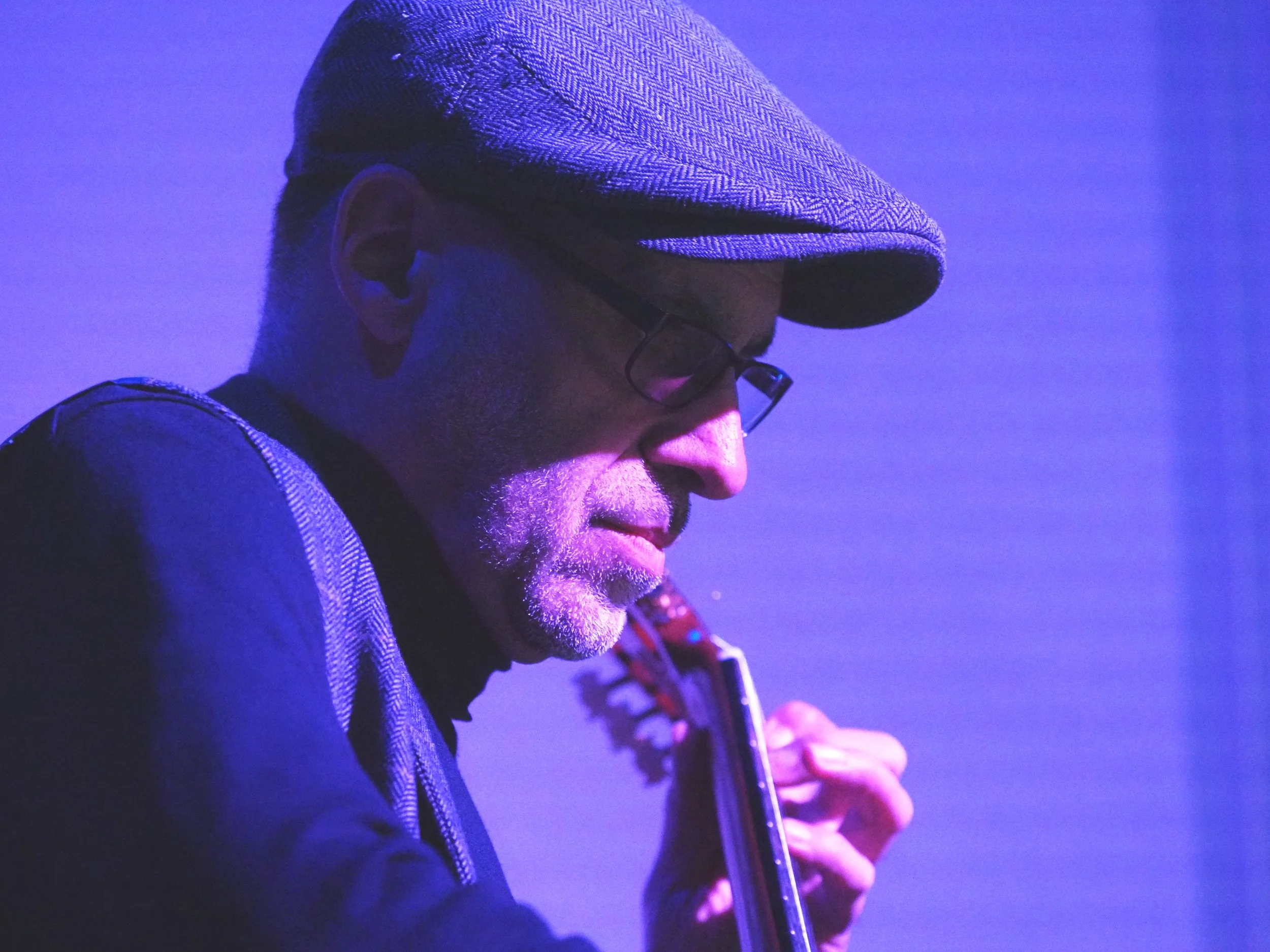 Ein Mann trägt eine Mütze und Brille, spielt Gitarre, in einer blau-violetten Beleuchtung.