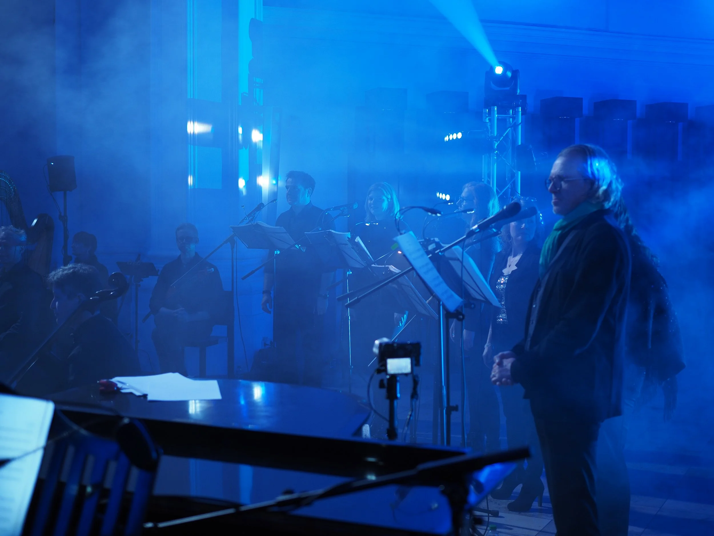 Mehrere Musiker stehen auf einer Bühne, umgeben von blauen Licht- und Nebeleffekten, während sie mit ihren Instrumenten spielen.