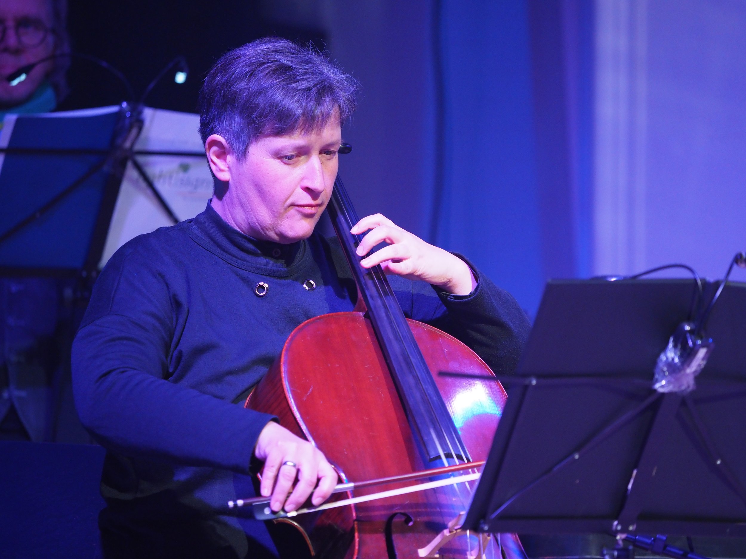 Ein Musiker spielt ein Cello auf einer Bühne, umgeben von blauen Lichteffekten.
