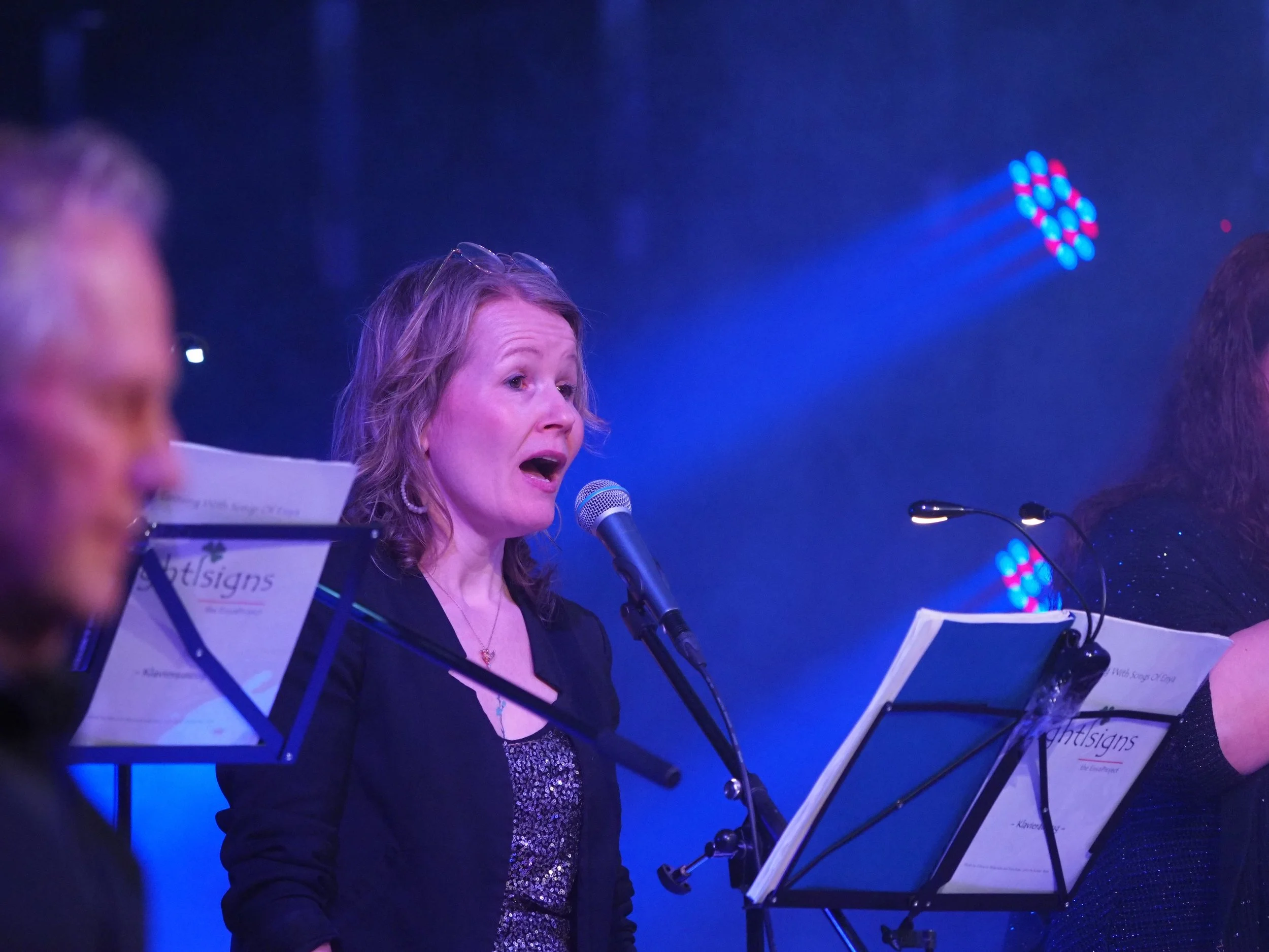 Frau singt bei einem Konzert, liest Noten, im Hintergrund bunte Lichteffekte.