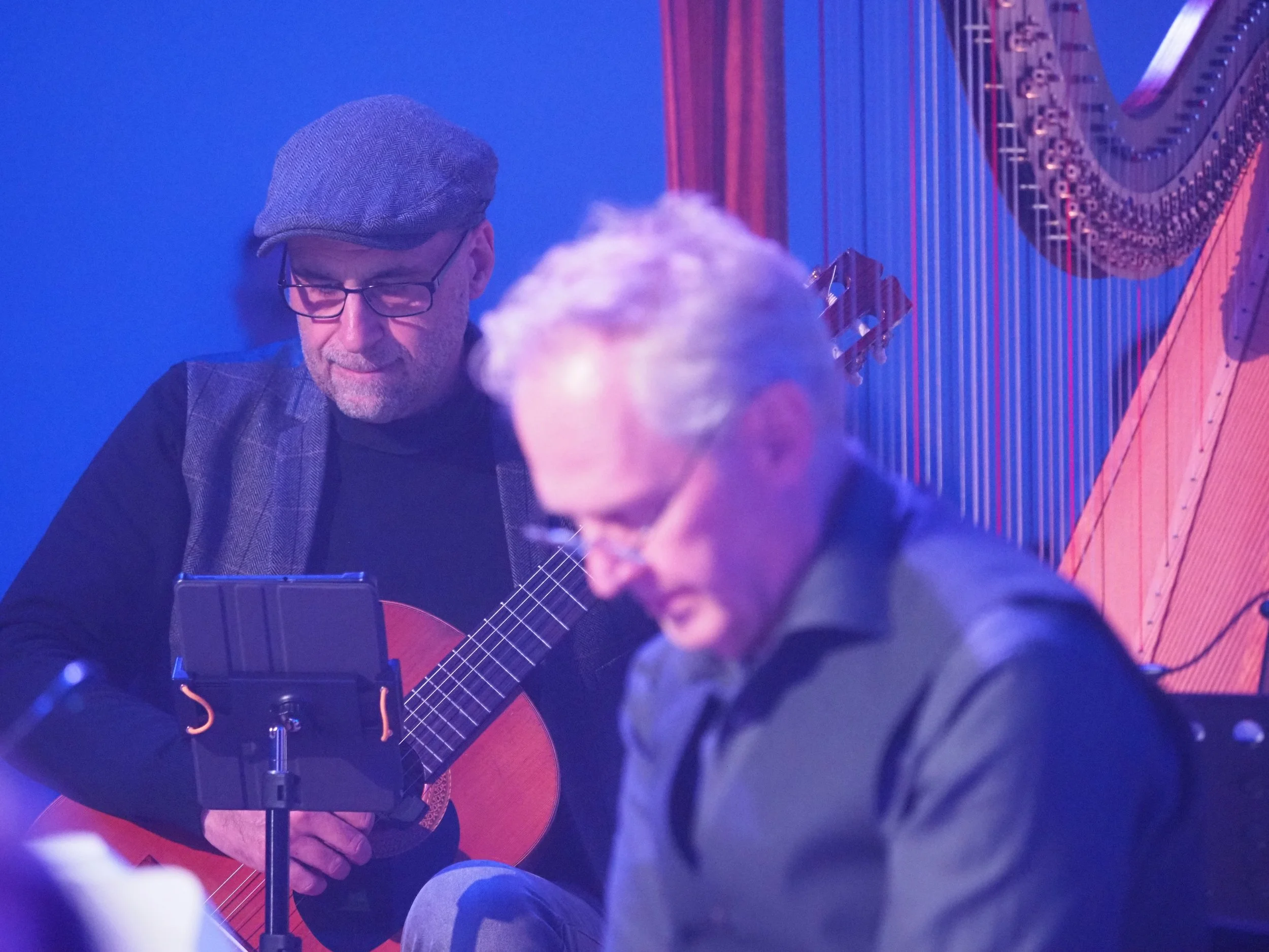 Zwei Männer, einer spielt Gitarre, während der andere in einem dunklen Raum mit blauer Beleuchtung sitzt.