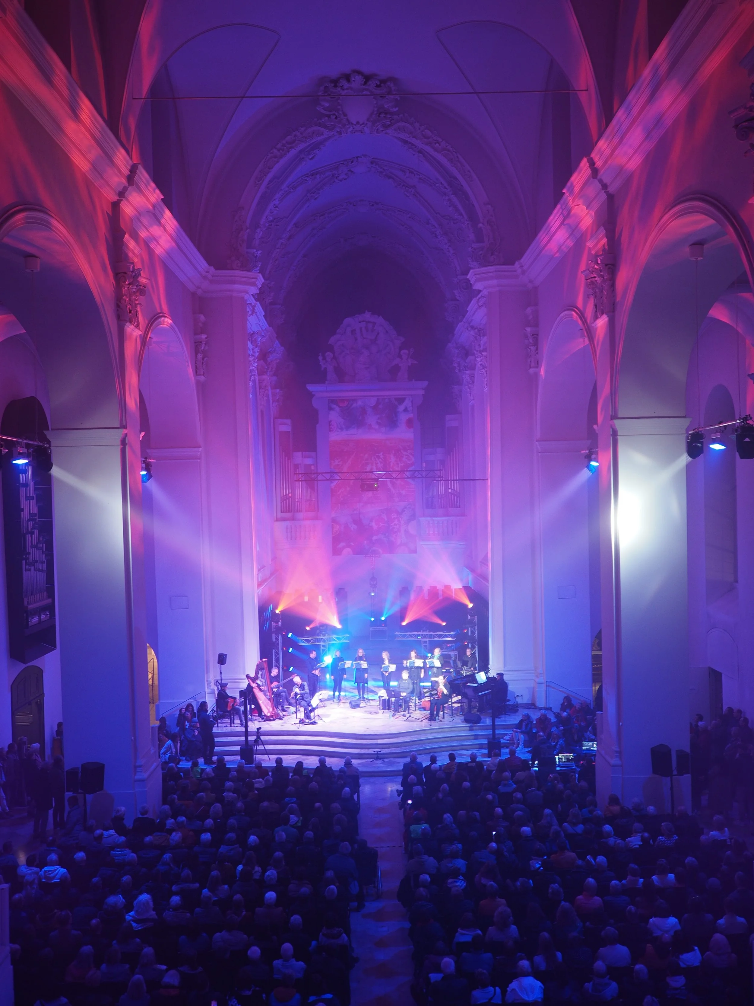 Ein Orchester spielt auf einer Bühne in einer prachtvollen, hochdekorierten Kirche bei einer musikalischen Veranstaltung. Das Publikum sitzt im dunklen Kirchenraum und schaut zu.