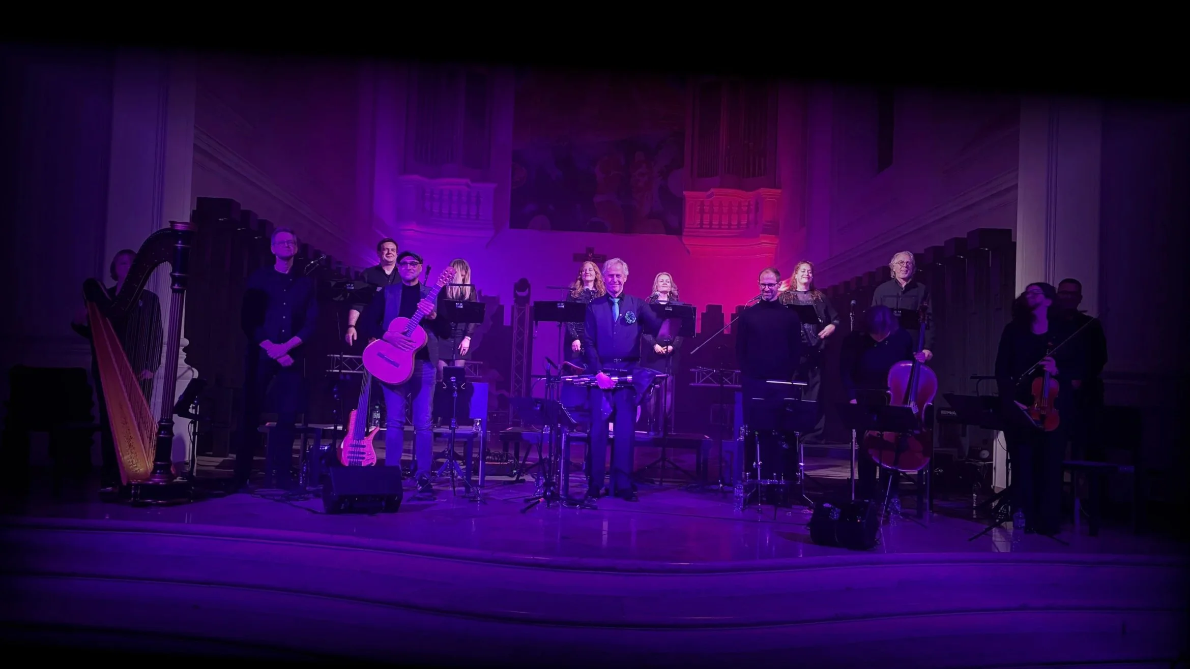 Eine Gruppe von Musikern auf einer Bühne, mit verschiedenen Instrumenten, im Konzertsaal mit pinker Beleuchtung.