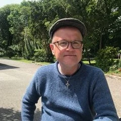 Ein Mann mit Brille, grauem Hut und blauer Jacke steht im Freien vor einer grünen, bewaldeten Umgebung.