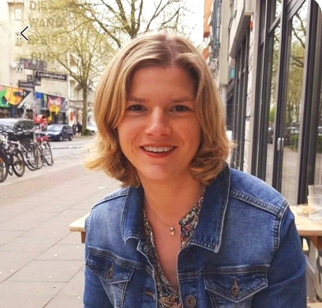 Junge Frau mit kurzen blondem Haar, lächelnd, trägt eine Jeansjacke, sitzt im Freien an einem Tisch in einer Stadtstraße mit Bäumen, Fahrrädern und Geschäften im Hintergrund.