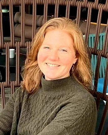Porträt einer lächelnden Frau mit roten Haaren, die einen grünen Pullover trägt.