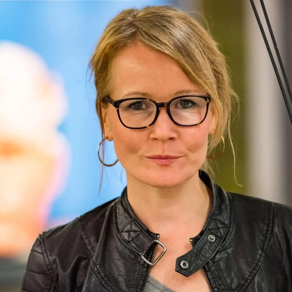Eine Frau mit kurzen, blonden Haaren, trägt eine schwarze Lederjacke und große runde Brillen.