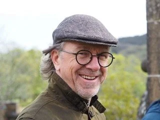 Ein lächelnder Mann mit grauem Haar, Bart, Brille, Mütze und dunkler Jacke im Freien mit Bäumen im Hintergrund.