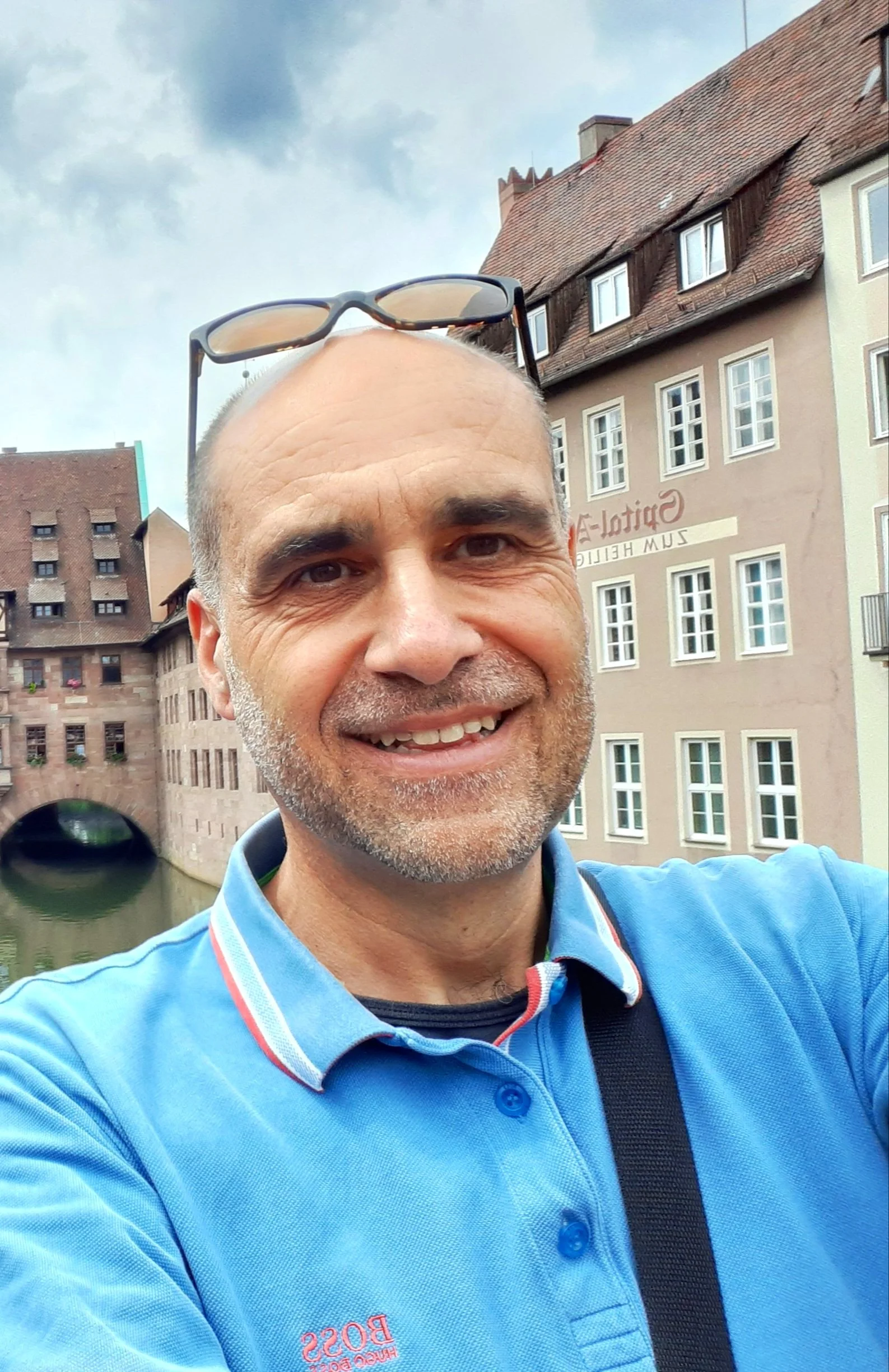 Nahaufnahme eines lächelnden Mannes mit Sonnenbrille auf dem Kopf, im Hintergrund ein Fluss, alte Gebäude und eine Brücke in einer Stadtumgebung.