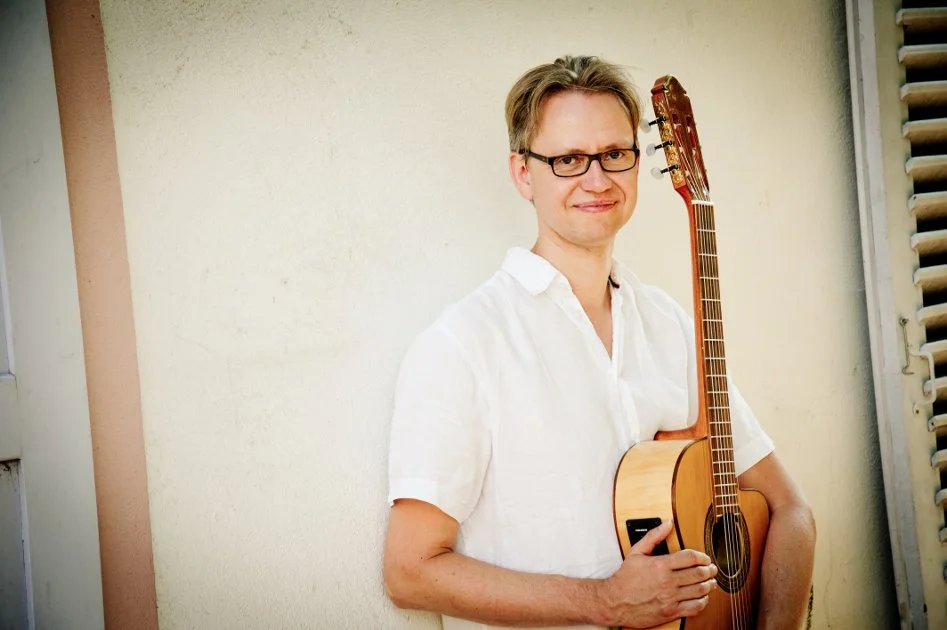 Mann mit Brille und weißen Hemd hält eine Gitarre vor einer hellen Wand.