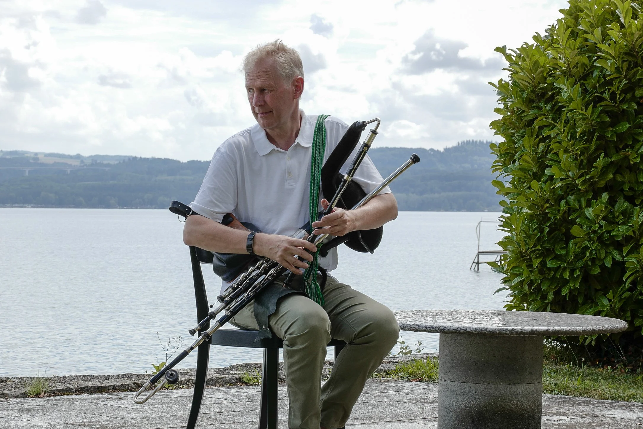 Ein älterer Mann sitzt auf einer Bank am Wasser und spielt Ulliaen Pipes