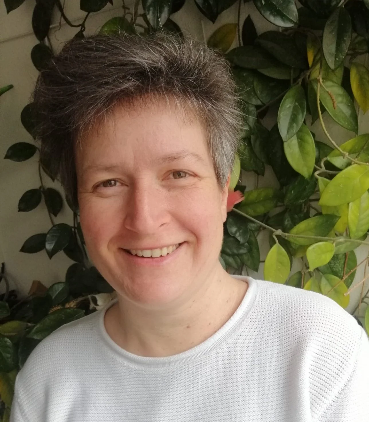 Lächelnde Frau mit kurzen grauen Haaren vor einer grünen Pflanzenwand.