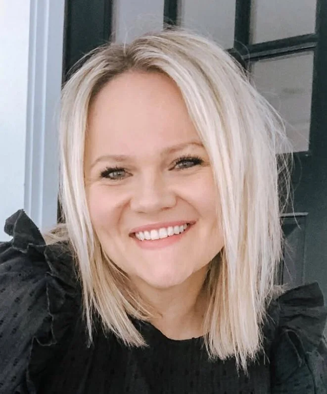 Close-up photo of a smiling woman with shoulder-length blonde hair wearing a black top, indoors with a dark background.