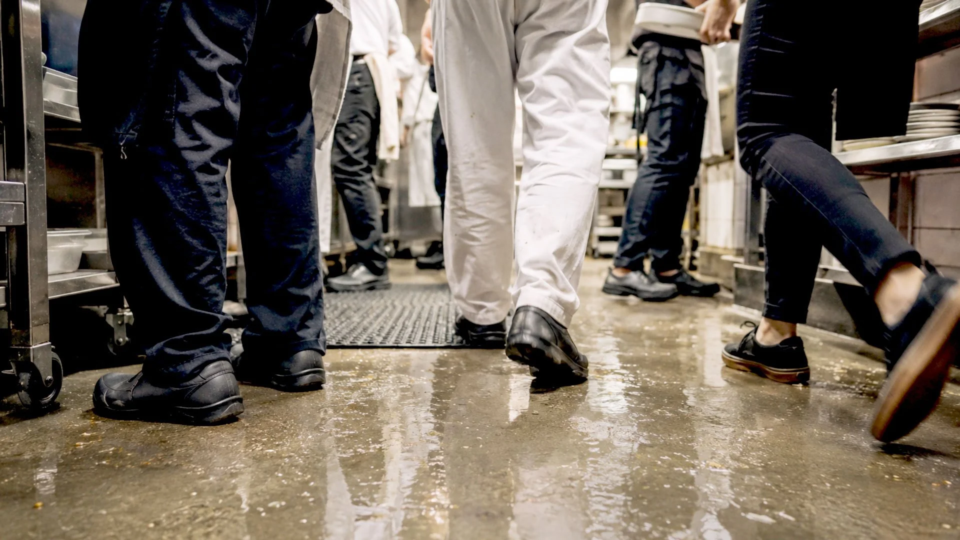 People wearing black shoes and pants, and one person in white scrubs, standing on a wet, dirty kitchen floor in a restaurant kitchen.