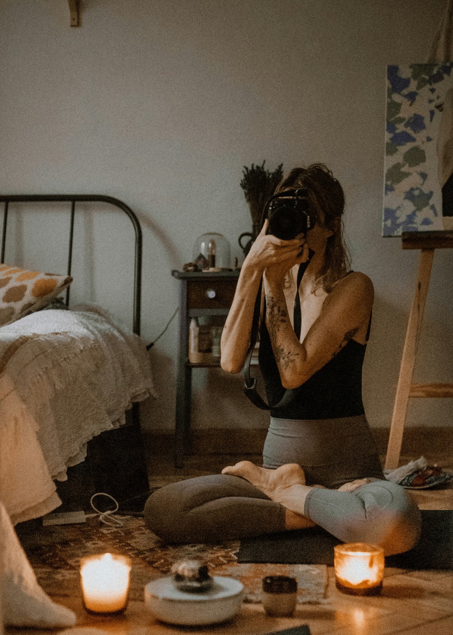 A woman with tattoos on her arm practicing yoga on a mat in a cozy, dimly-lit room, taking a photo of herself with a camera. Candles and a bowl with stones are on the floor in front of her, and a bed with patterned pillows and a nightstand with decorative items are in the background.