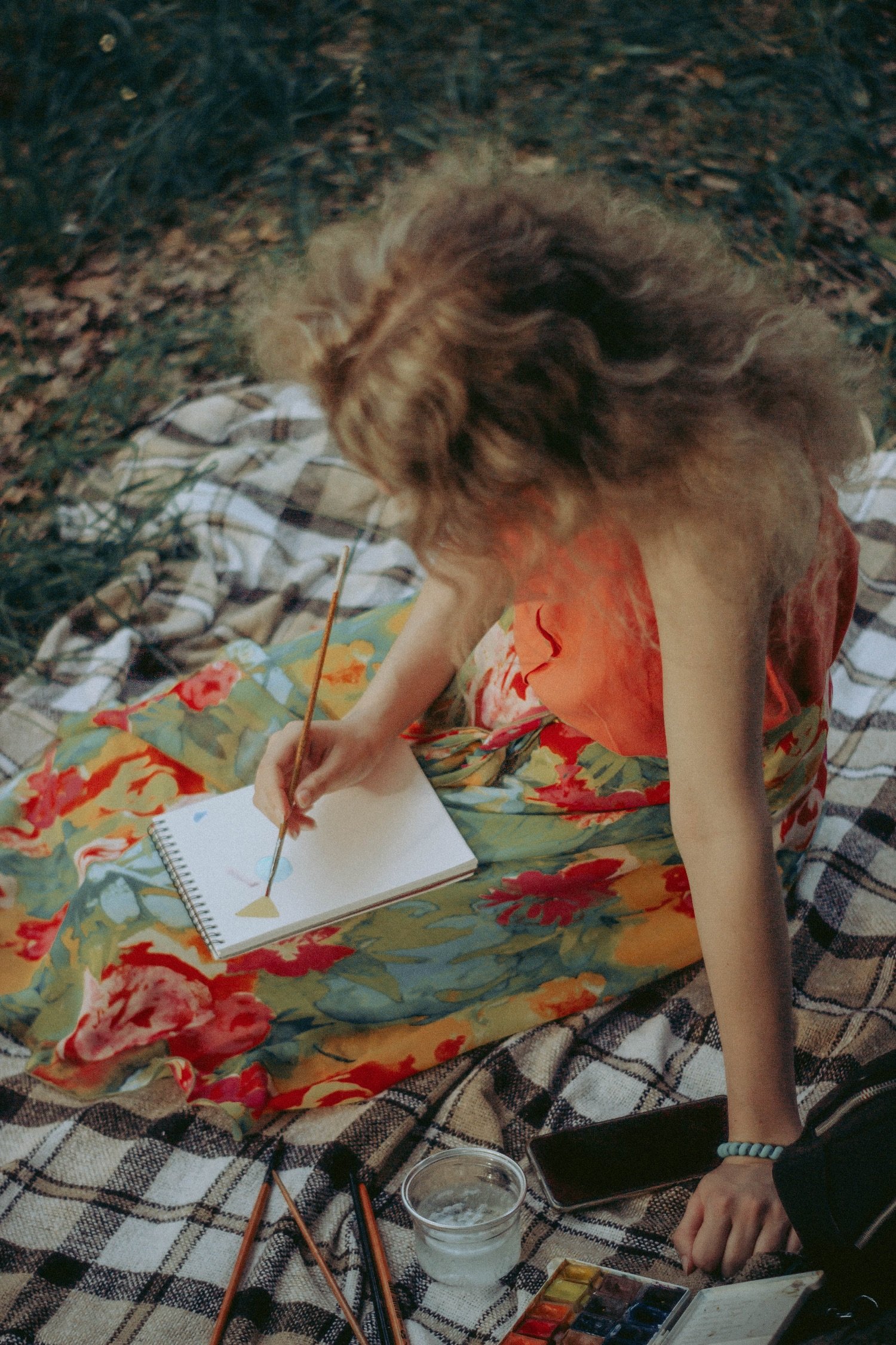 Girl in a floral dress sitting on a blanket outdoors, painting in a sketchbook, with paint supplies nearby.