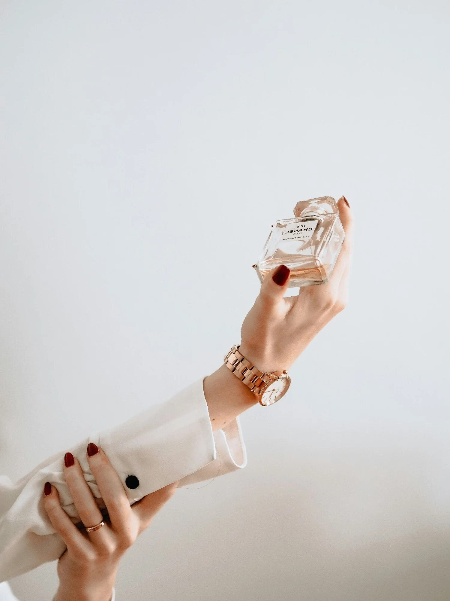Person holding a Chanel No. 5 perfume bottle with a white background, wearing a white shirt, gold watch, and red nail polish.
