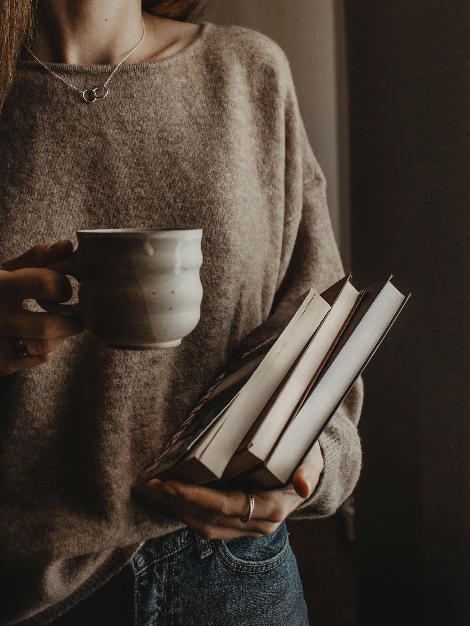 Person wearing a beige sweater, holding a white mug in one hand and three books in the other.