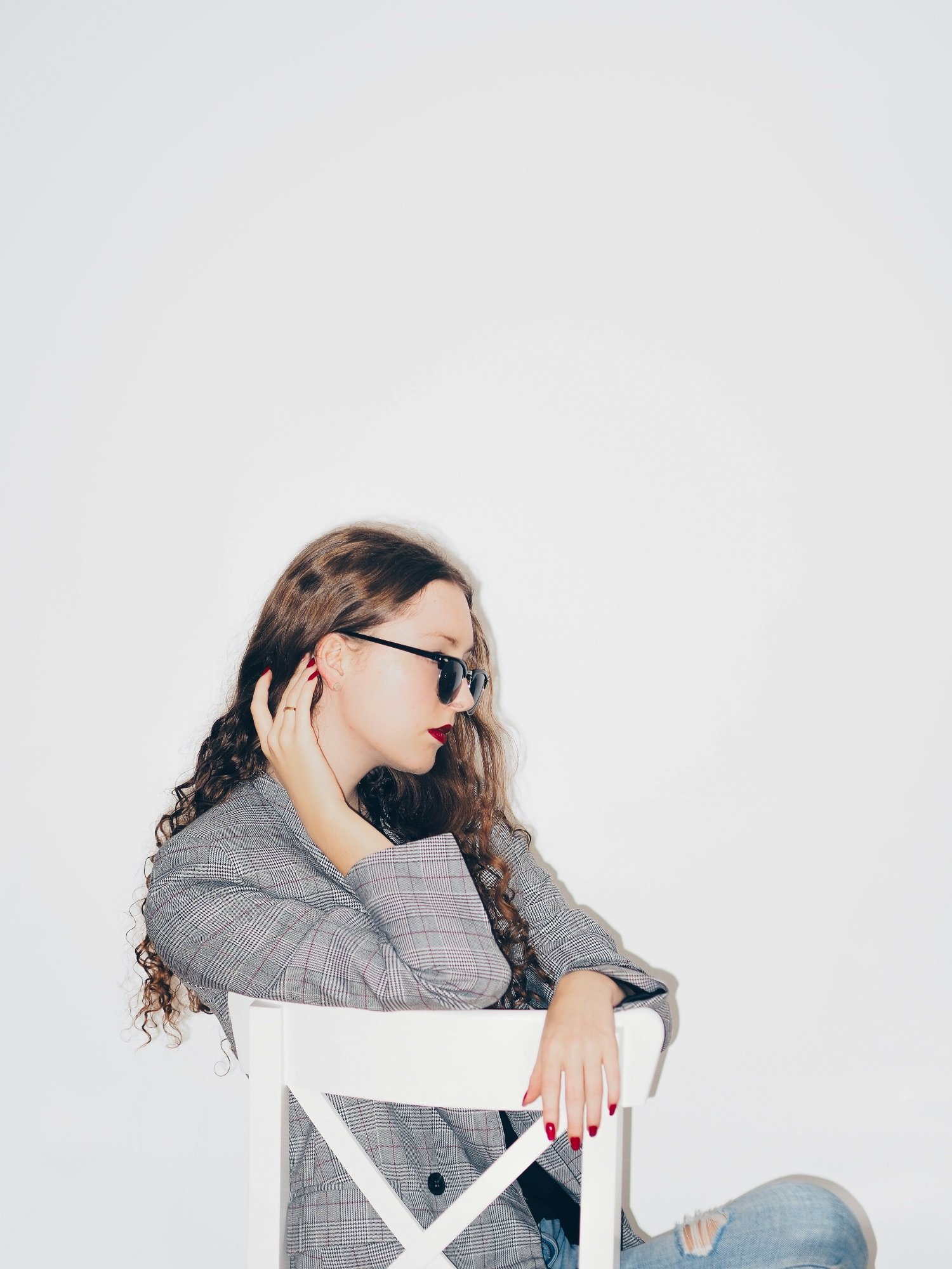 A woman with curly hair wearing sunglasses, a gray plaid blazer, and red lipstick sitting on a white chair against a plain white background, looking downward with her hand touching her ear.