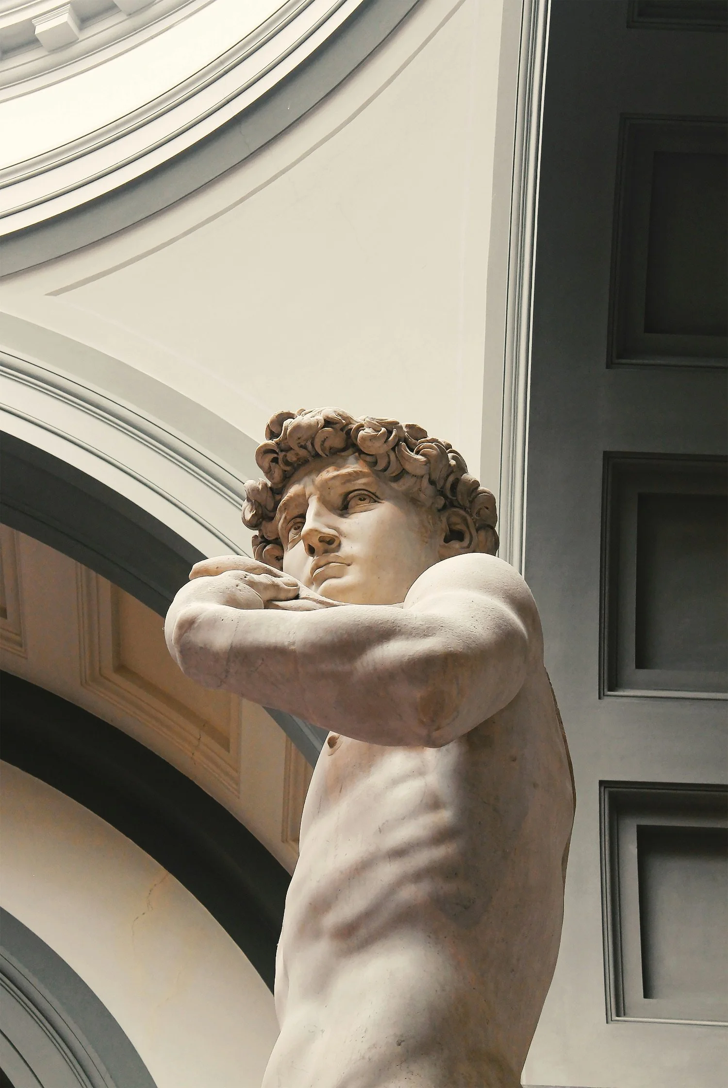 Close-up view of a classical marble sculpture of a male figure with curly hair, shown from the waist up, with arms crossed over the chest, inside a building with arched ceilings and decorative moldings.