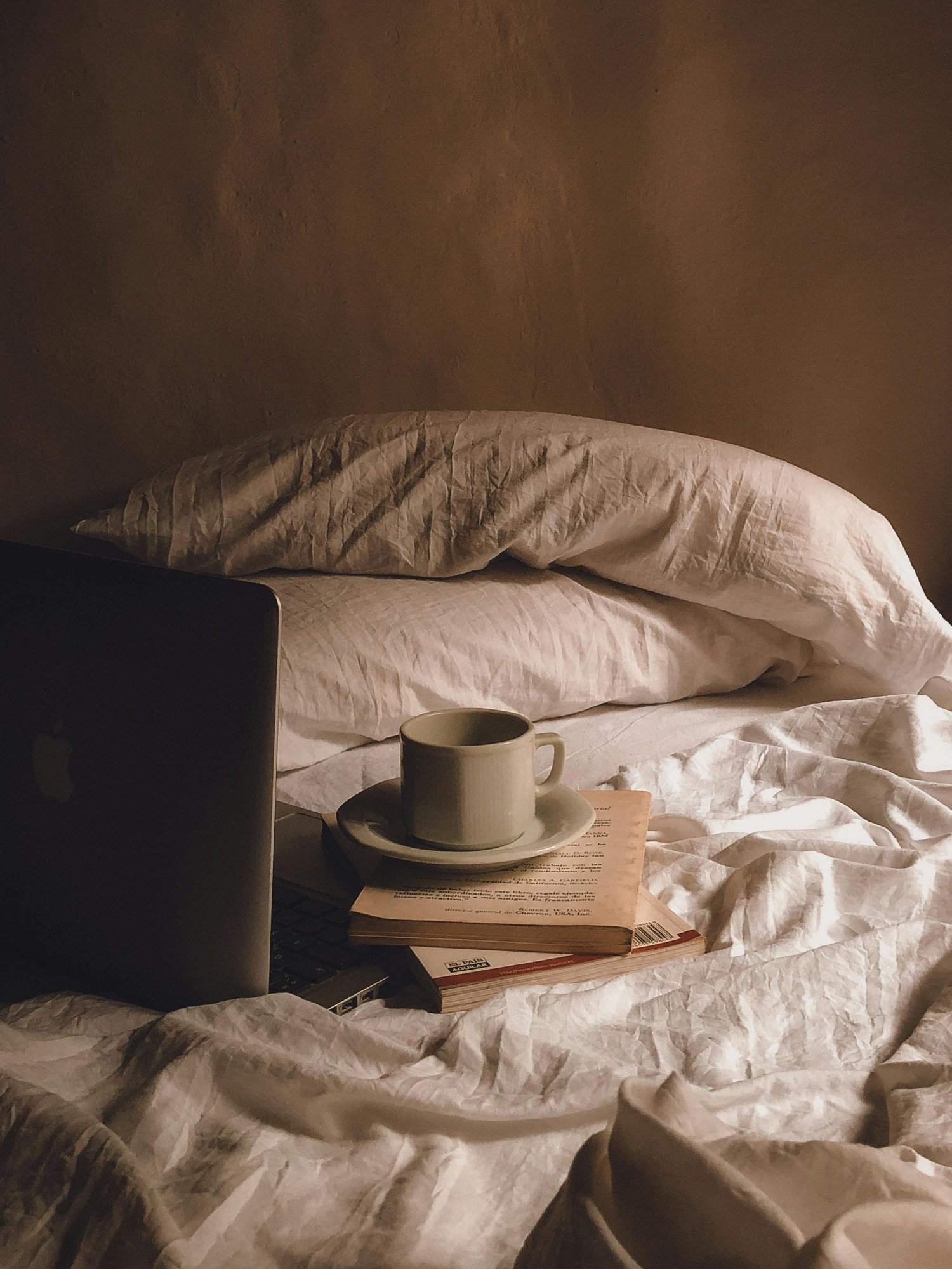 Bed with crumpled white sheets, a stack of books, a white coffee cup on a saucer, and a laptop on a bedside table.