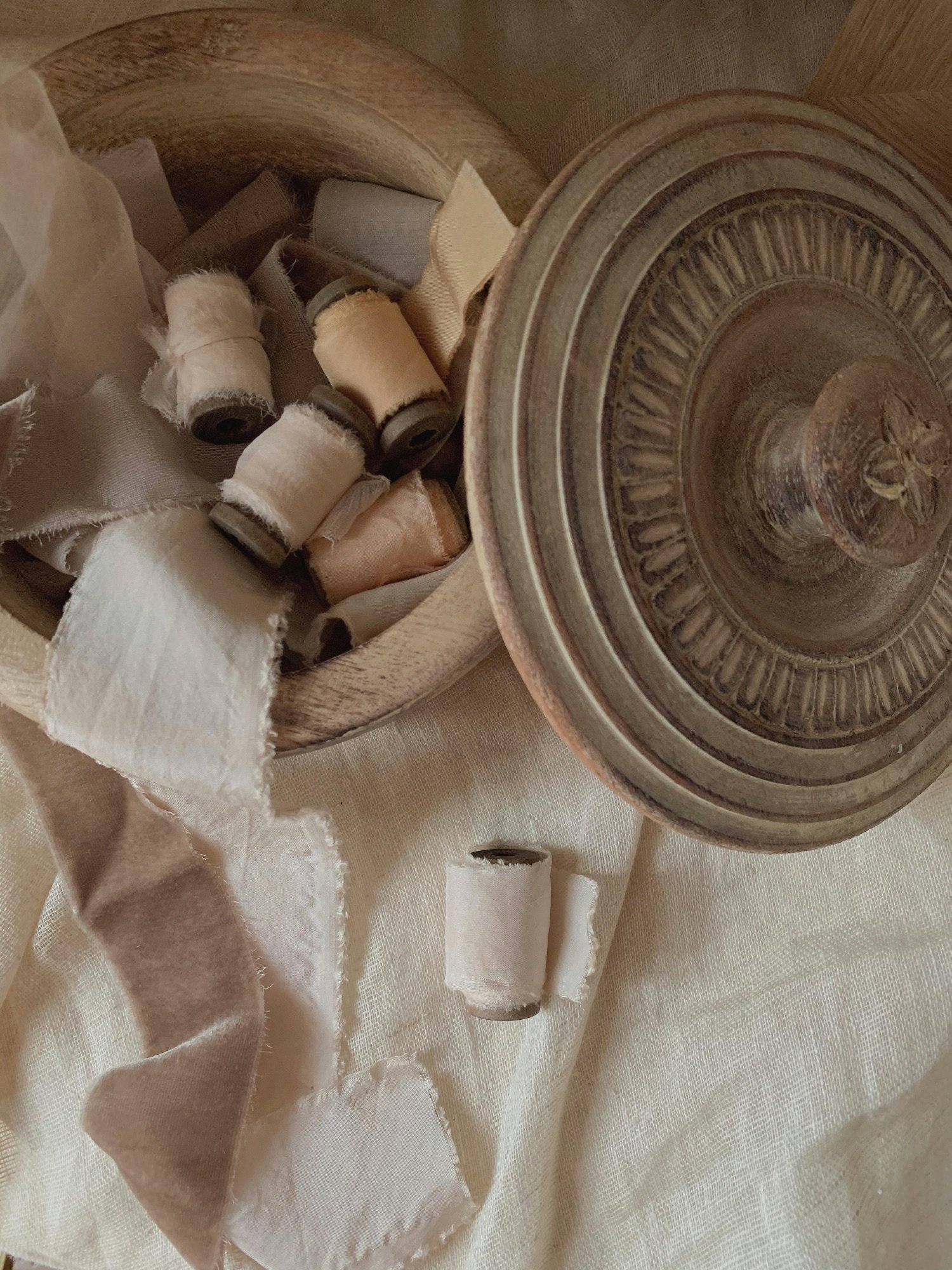 A wooden bowl filled with spools of beige, white, and cream fabric and ribbon, with some loose pieces and a decorative glass bowl nearby on a light-colored fabric surface.