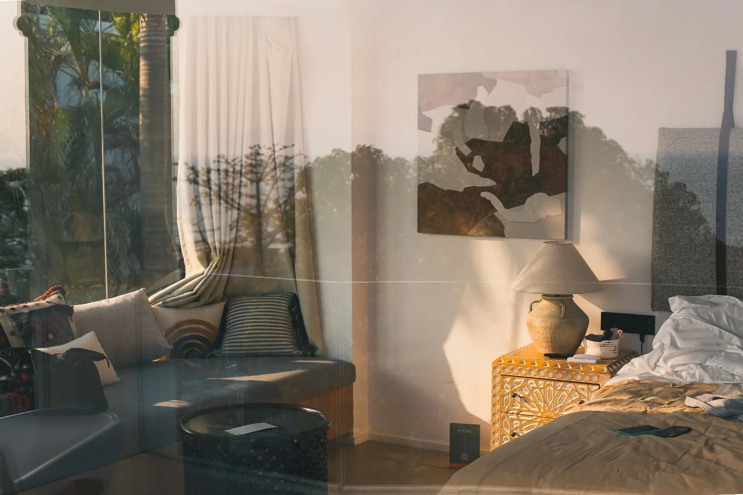 A bedroom with a bed, a nightstand with a lamp, and a window with curtain and a seating area with pillows, reflected through glass.
