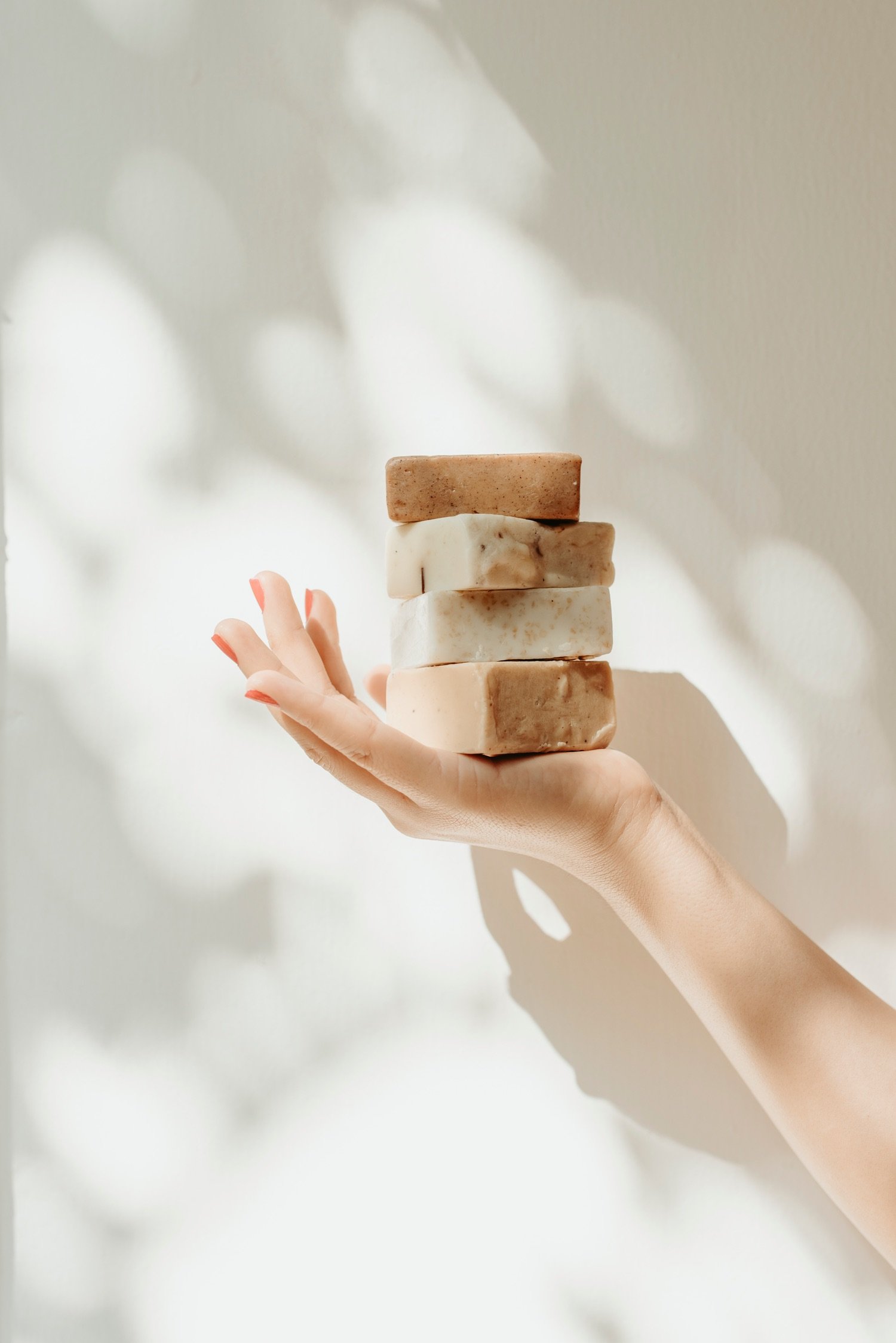 A hand holding a stack of five different soap bars against a light background with soft, out-of-focus light patterns.